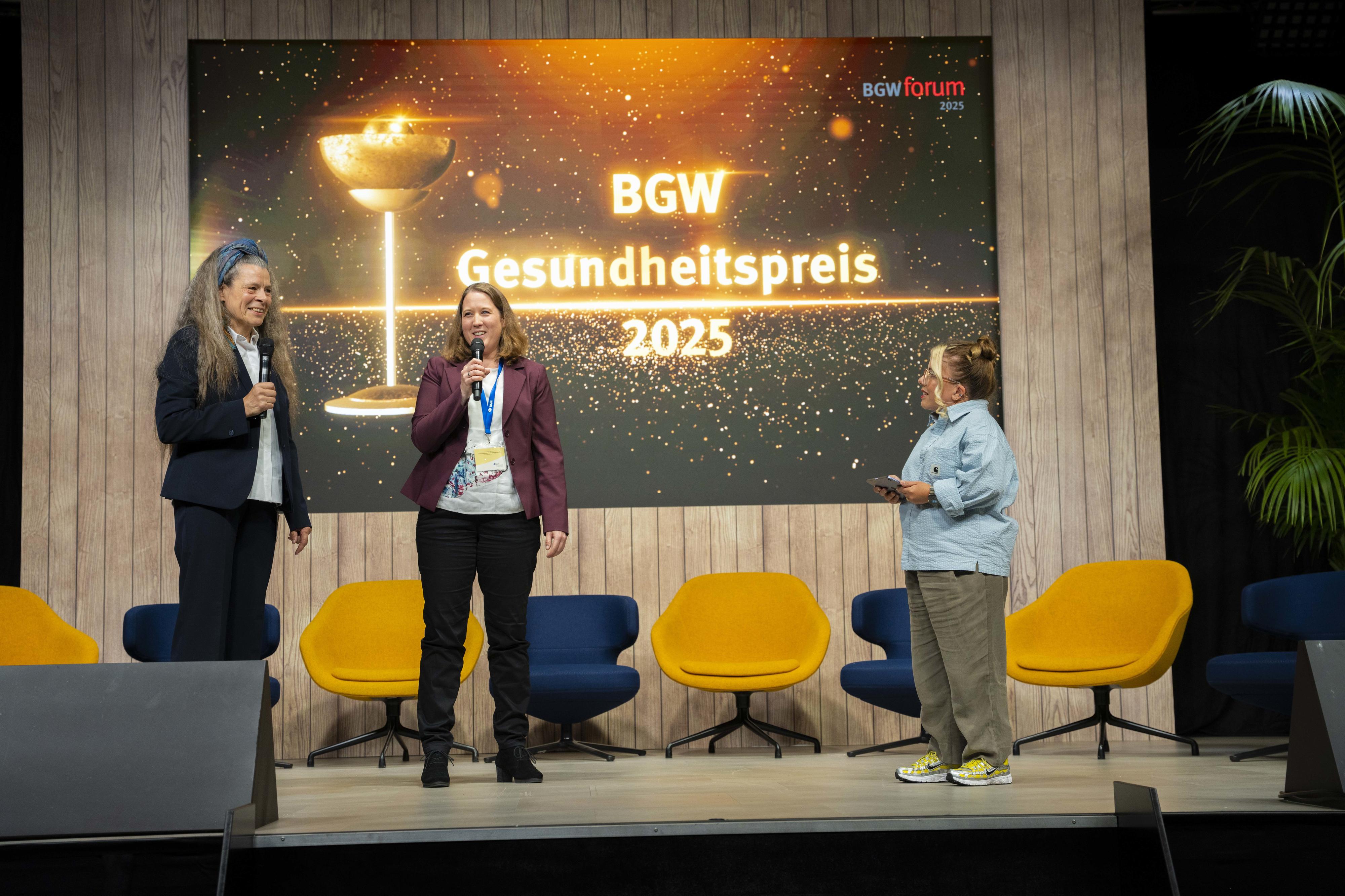 Drei Personen auf einer Bühne, eine davon hält sich ein Mikrofon an den Mund
