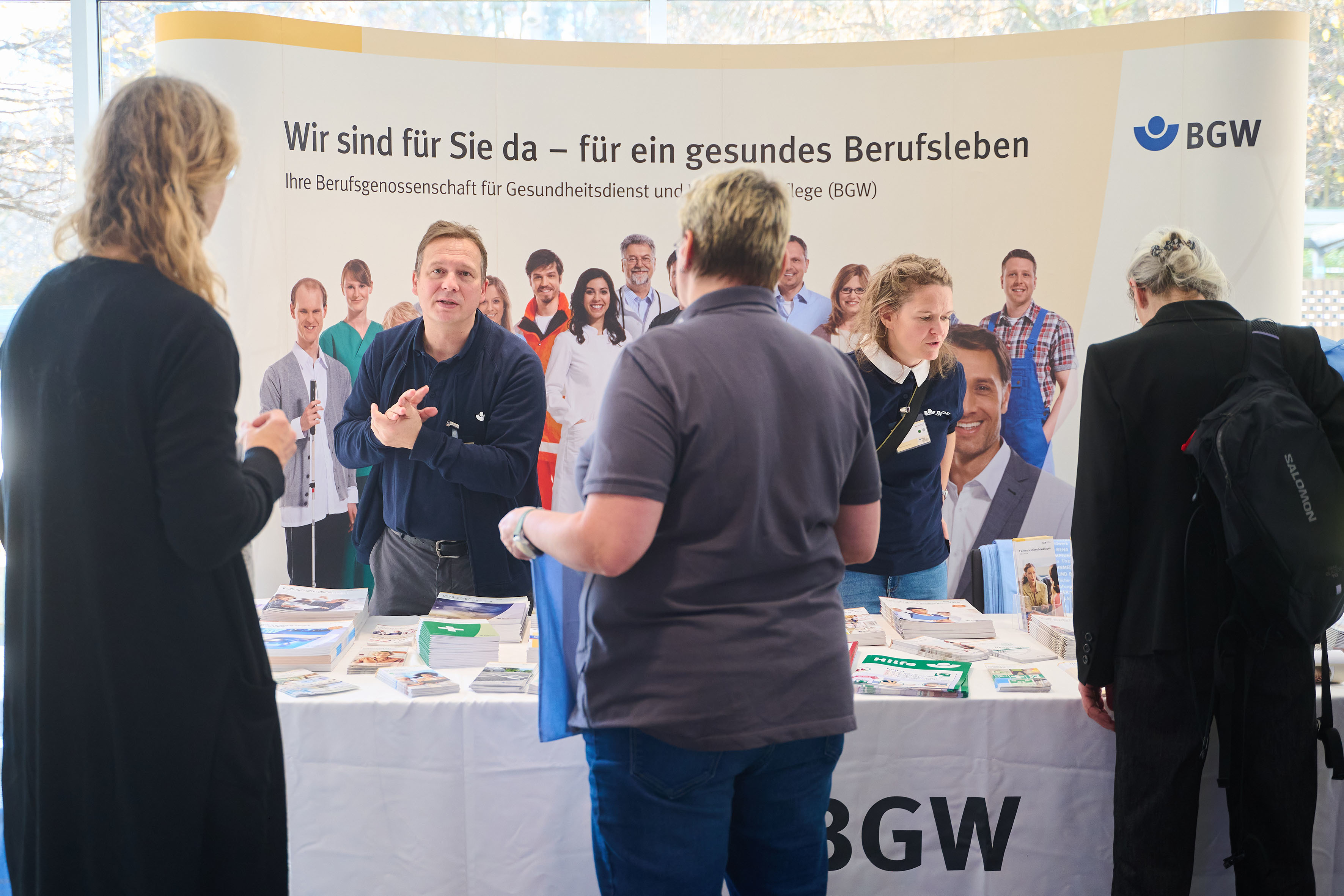 Menschen informieren sich an einem Messestand der BGW, es liegen Broschüren aus.