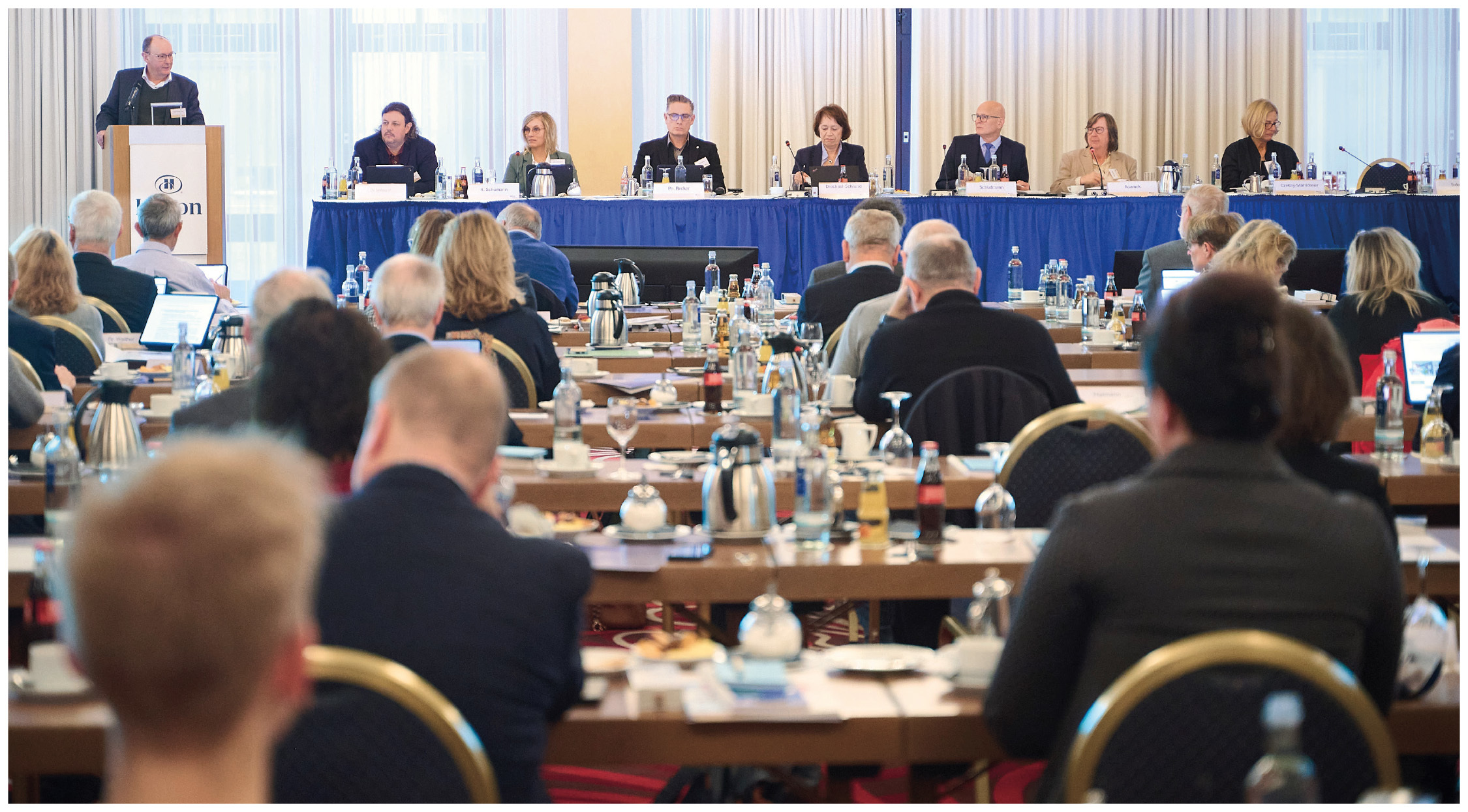 Ein Saal, an der Stirnseite ein Pult und ein langer Tisch an denen Personen sitzen, vor ihnen ein Publikum