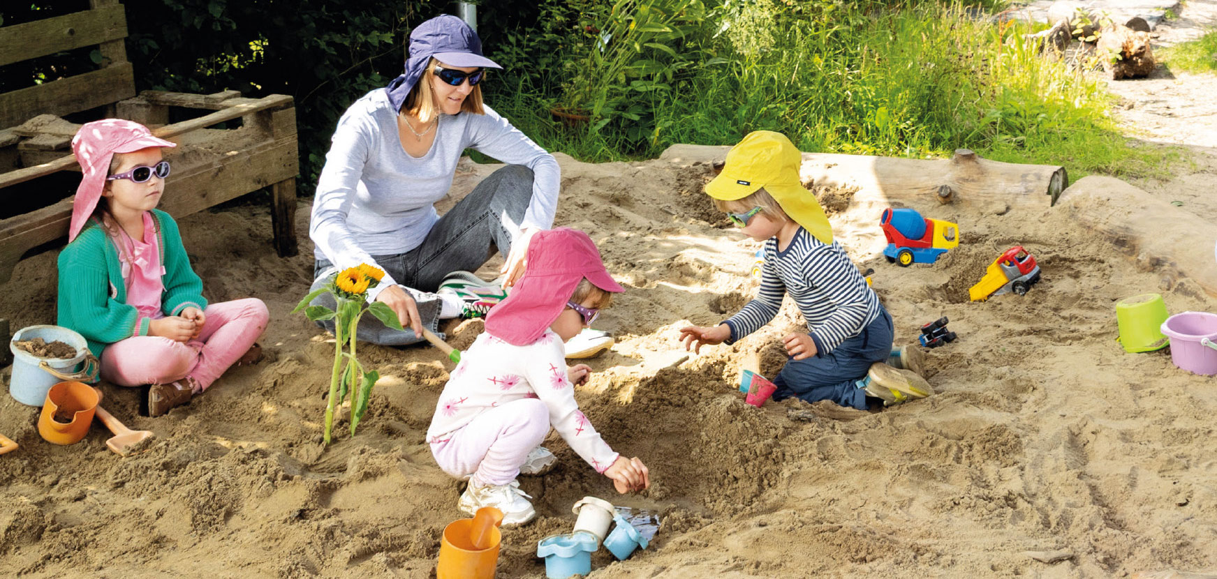 Drei Kinder und eine Erwachsene mit Sonnenbrill und Hut mit Nackenschutz in einem Sandkasten, im Sand liegen Spielgeräte.