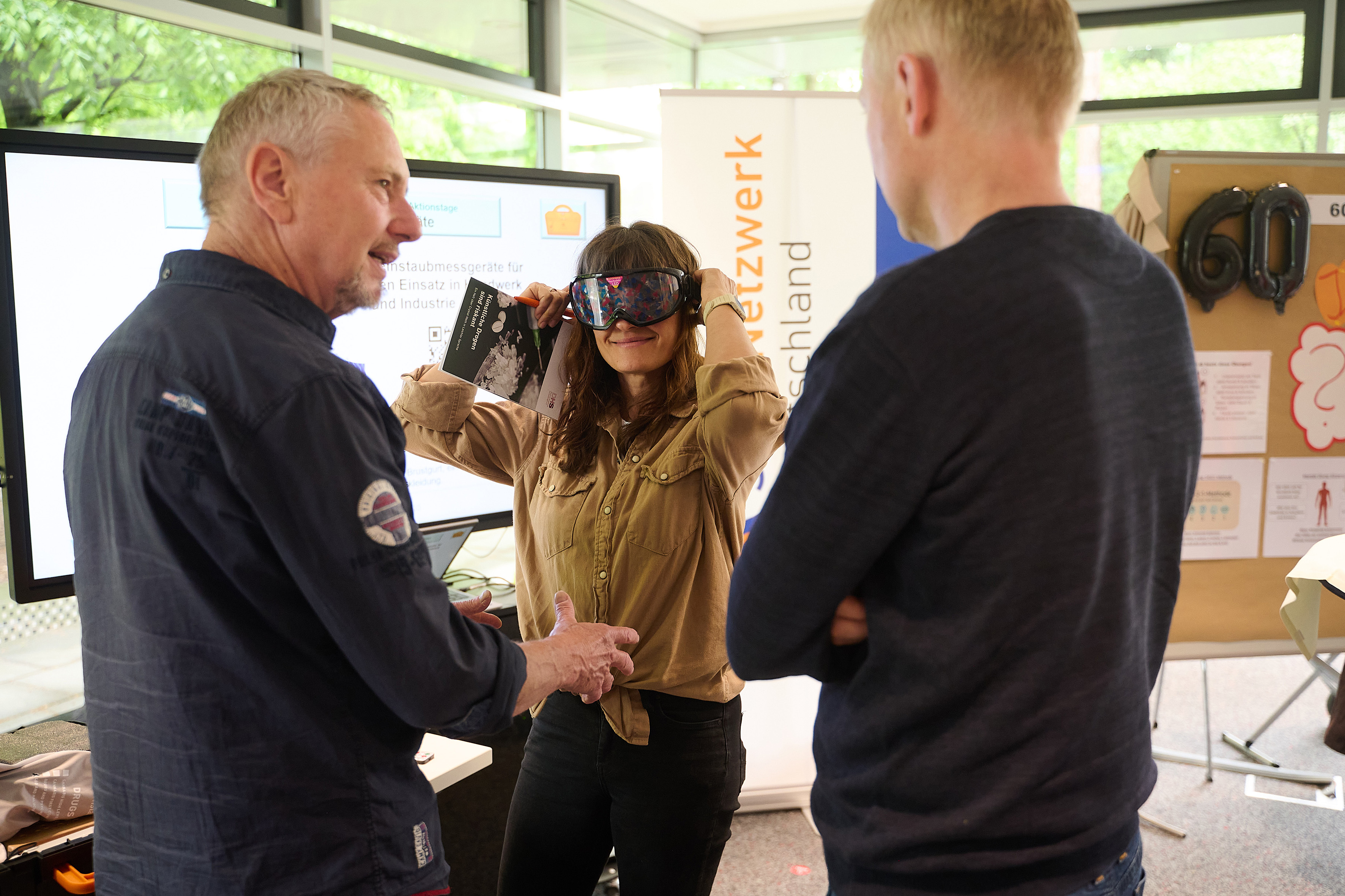 Eine Frau setzt eine VR-Brille auf, während ein ihr gegenüber stehender Mann einem anderen Mann etwas erklärt.