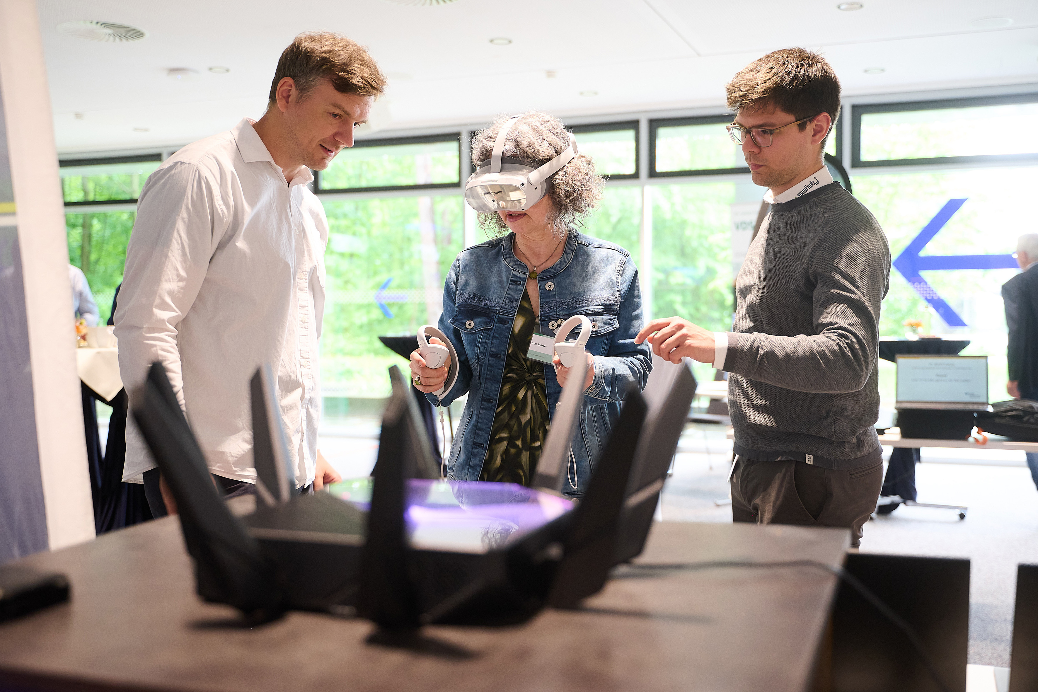 Eine Frau trägt eine VR-Brille auf dem Kopf und Controller in den Händen. Zwei Männer stehen neben ihr.