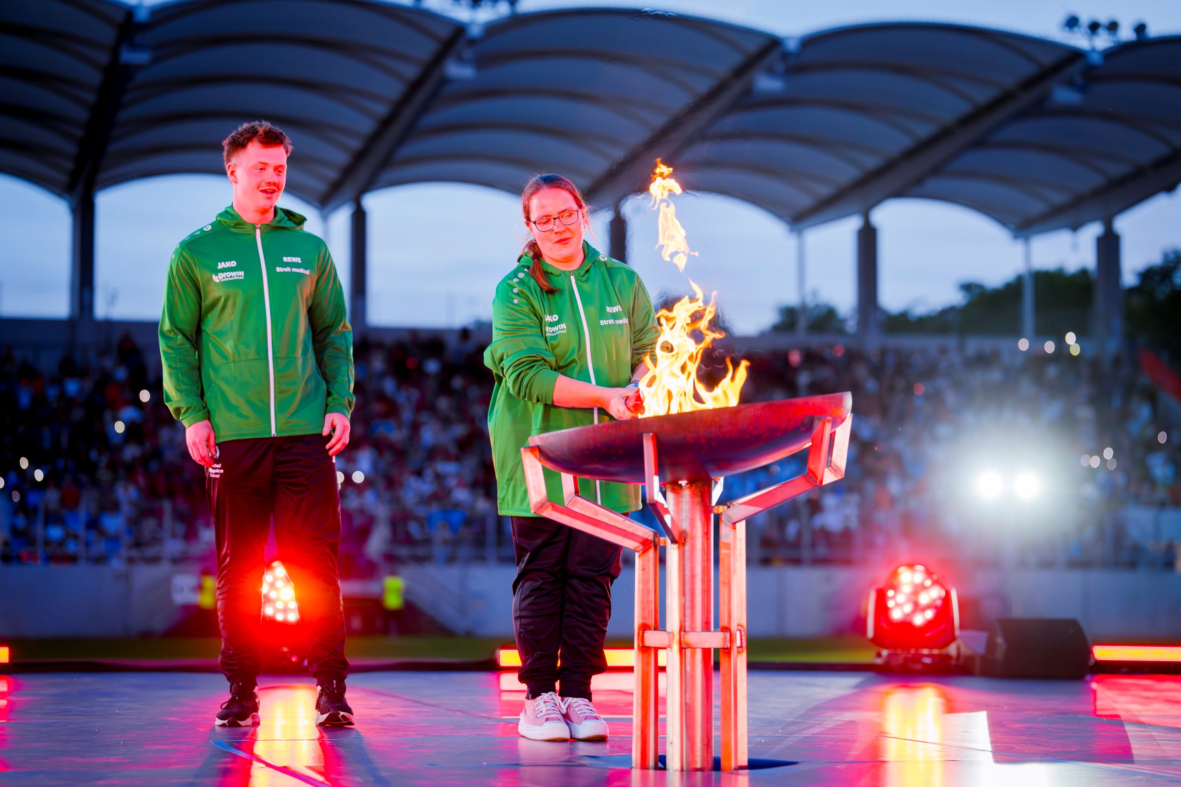 Ein Mann und eine Frau in einem Stadion. Die Frau entzündet mit einer Fackel das olympische Feuer in einer Feuerschale.