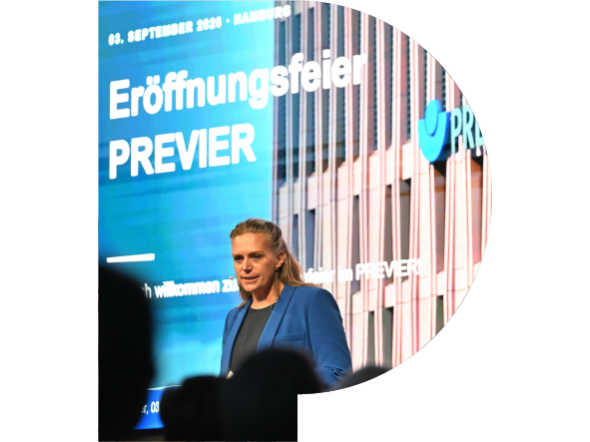 In dem PREVIER-P ist ein Foto von der Eröffnungsfeier des PREVIER. Die Moderatorin steht auf der Bühne vor einer Leinwand, auf dem ein Bild des PREVIER und die Worte "Eröffnungsfeier PREVIER" zu sehen sind.
