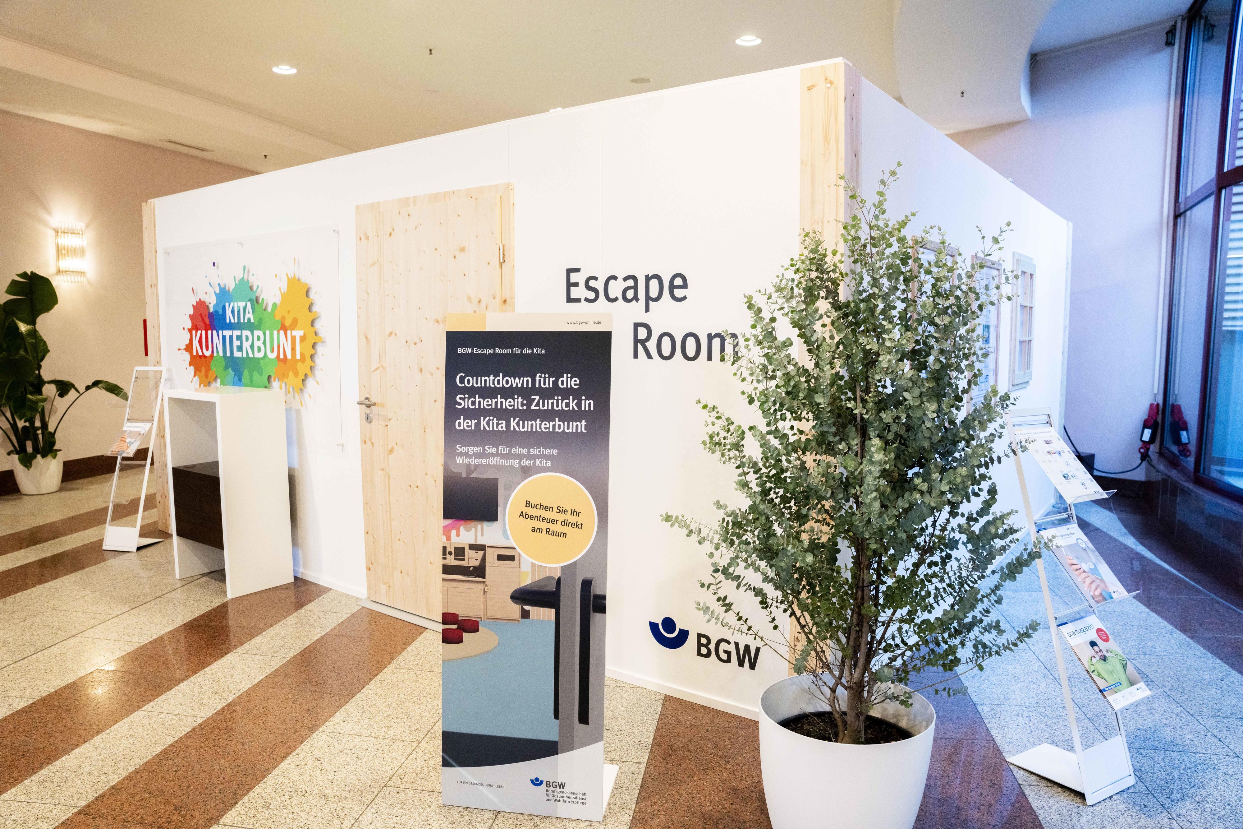 BGW-Messehäuschen mit Holztür, auf der Vorderseite des Häuschens prangen zwei Tafeln. Auf einer steht "Escape Room", auf einer anderen "Kita Kunterbunt". Das Häuschen hat eine Tür und mehrere Sprossenfenster, im Vordergrund ein Aufsteller "Countdown für die Sicherheit: Zurück in der Kita Kunterbunt. Sorgen Sie für eine sichere Wiedereröffnung der Kita. Buchen Sie Ihr Abenteuer direkt am Raum".
