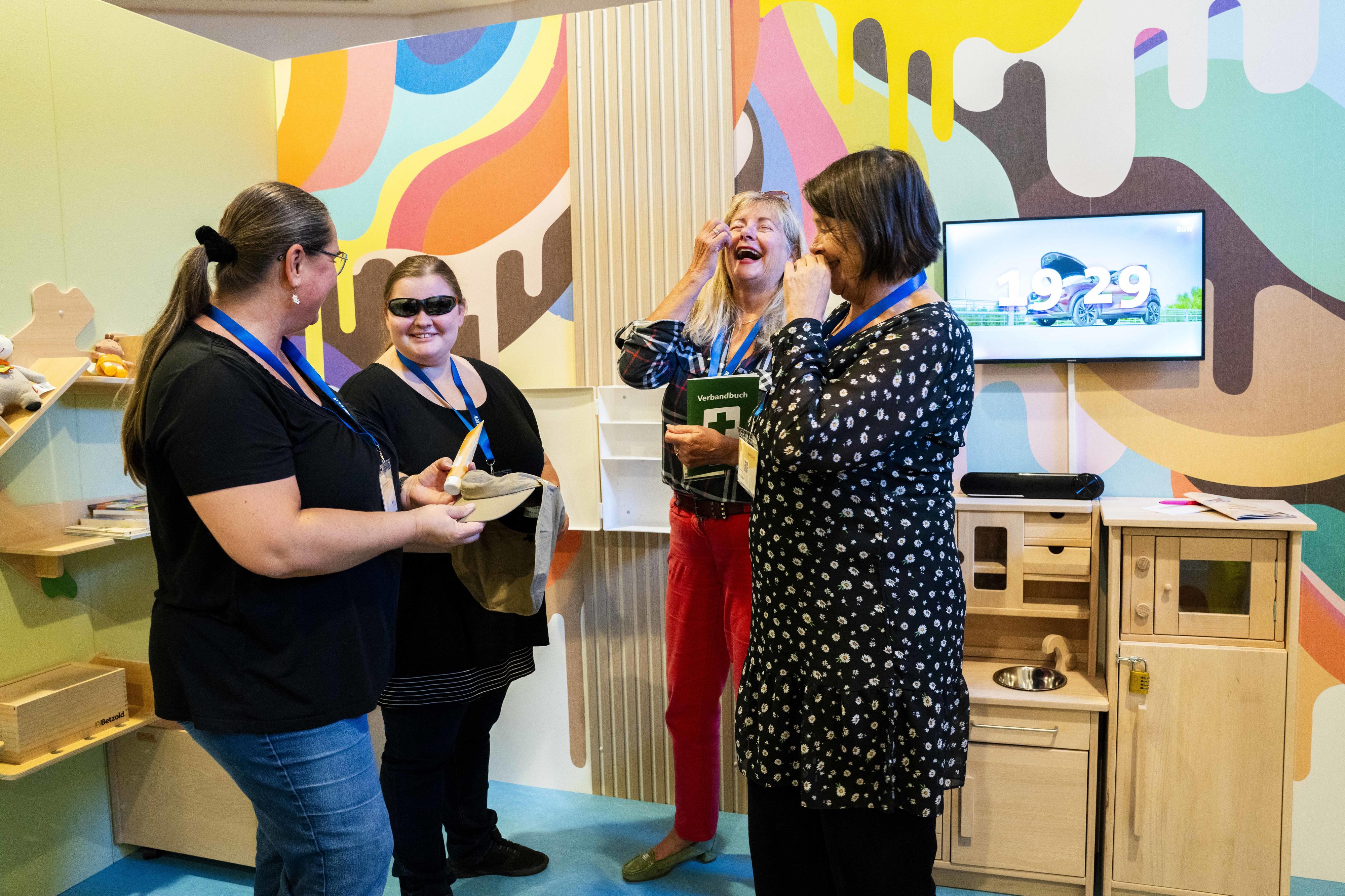 Vier Frauen im Gespräch, alle lachen amüsiert. Die Wand im Hintergrund ist mit bunten Farbklecksen geschmückt, hinten rechts ein hölzernes Regal mit Spielzeug und Büchern, außerdem eine hölzerne Spielküche und ein Monitor.Eine der Frauen trägt eine Sonnenbrille, eine weitere hält eine Tube Sonnencreme sowie einen Hut mit Nackenschut in der Hand, eine weitere Frau ein Verbandbuch.