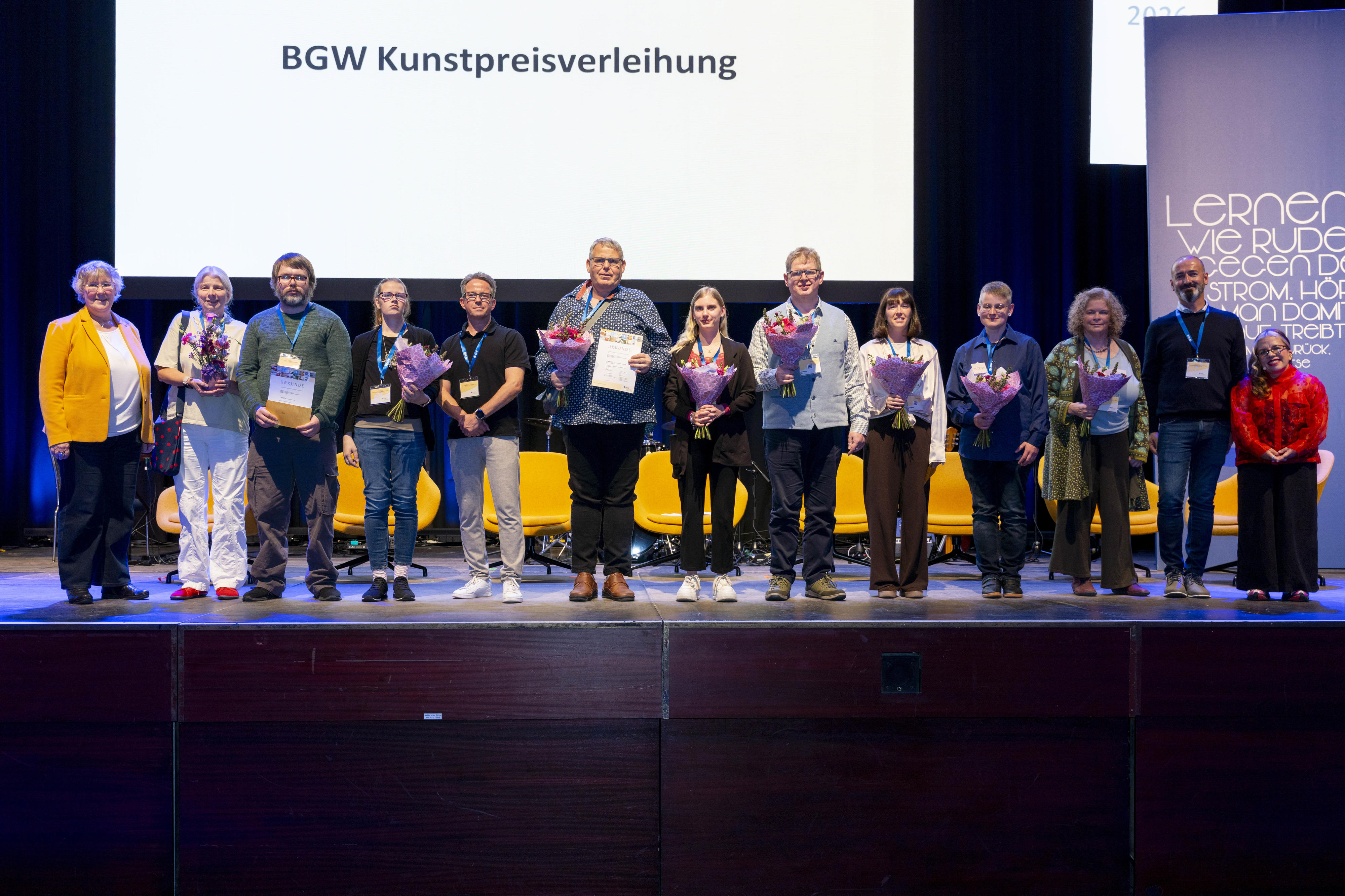 Dreizehn Personen stehen in einer Reihe auf einer Bühne, einige halten Blumen, andere Urkunden in den Händen. Im Hintergrund auf einer leuchtenden Leinwand steht "BGW Kunstpreisverleihung"