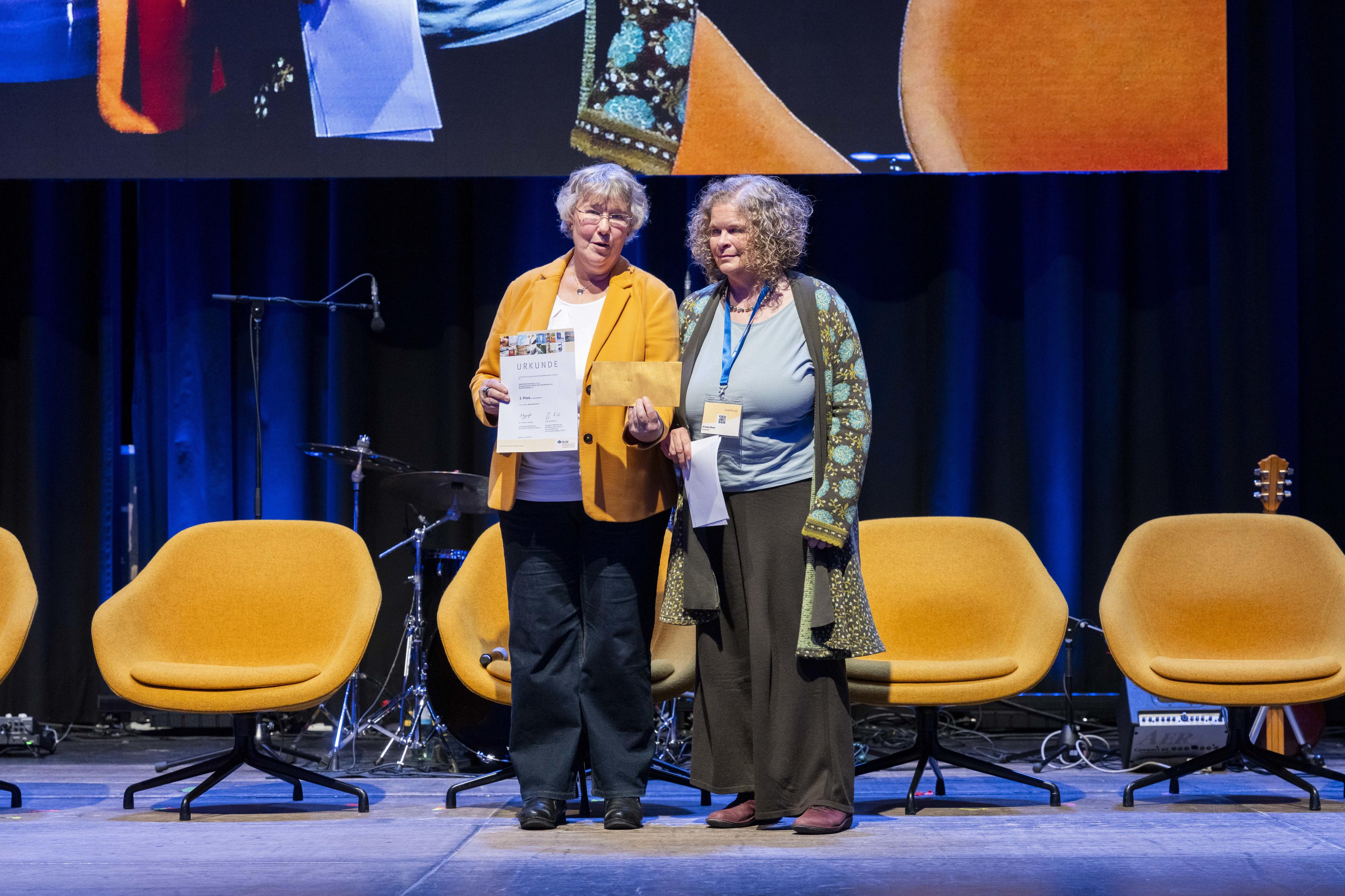 Silvia Thimm (l.) aus der BGW Selbstverwaltung mit der Gewinnerin des 3. Platzes Ursula Ebner auf einer Bühne.