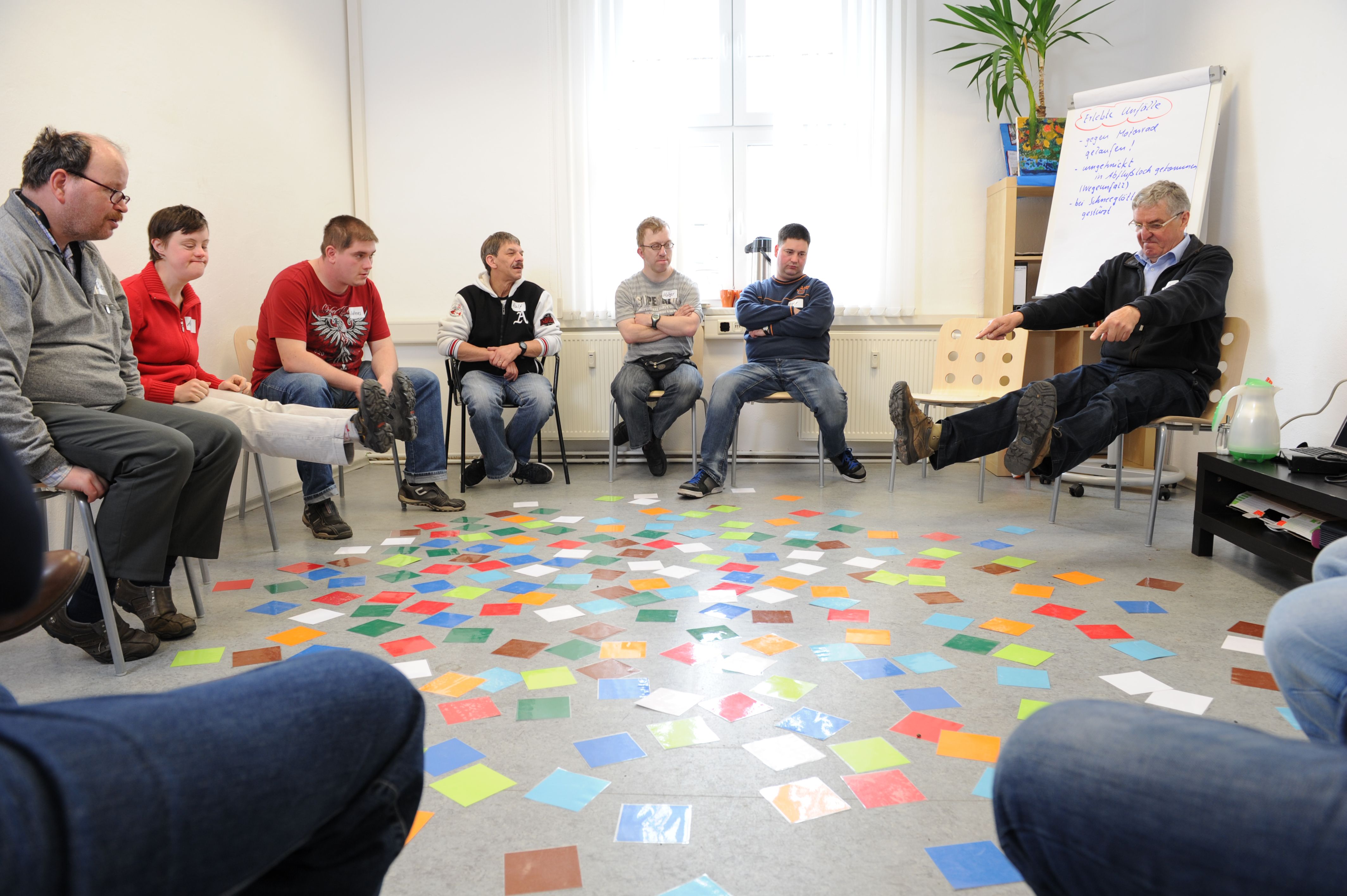 10 Personen sitzen im Kreis in einem Raum. Auf dem Boden liegen bunte Karten. Eine Person hebt die Beine und streckt die Arme nach vorne.
