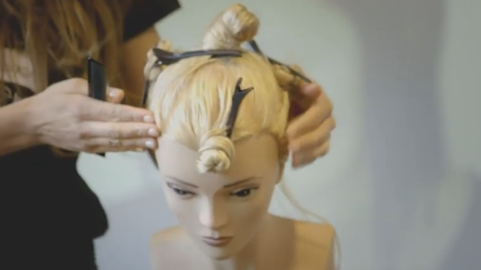 Frisierkopf mit blonden Haaren und Haarspangen wird von den beiden Händen einer Friseurin seitlich in Position gebracht