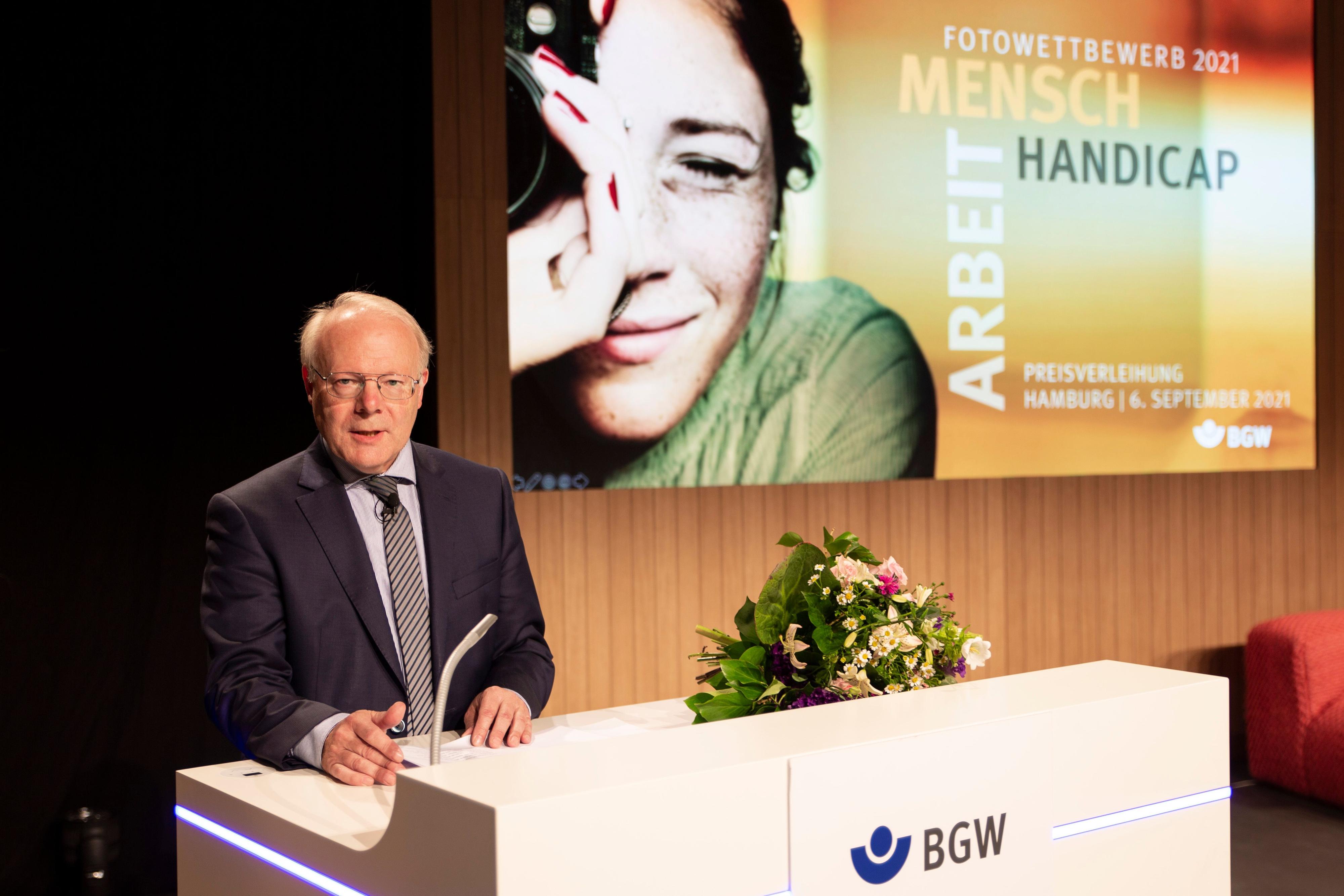 Prof. Dr. Stephan Brandenburg am Rednerpult, im Hintergrund Einblendung des Logos "Mensch - Arbeit - Handicap" und Bild einer Frau mit Kamera