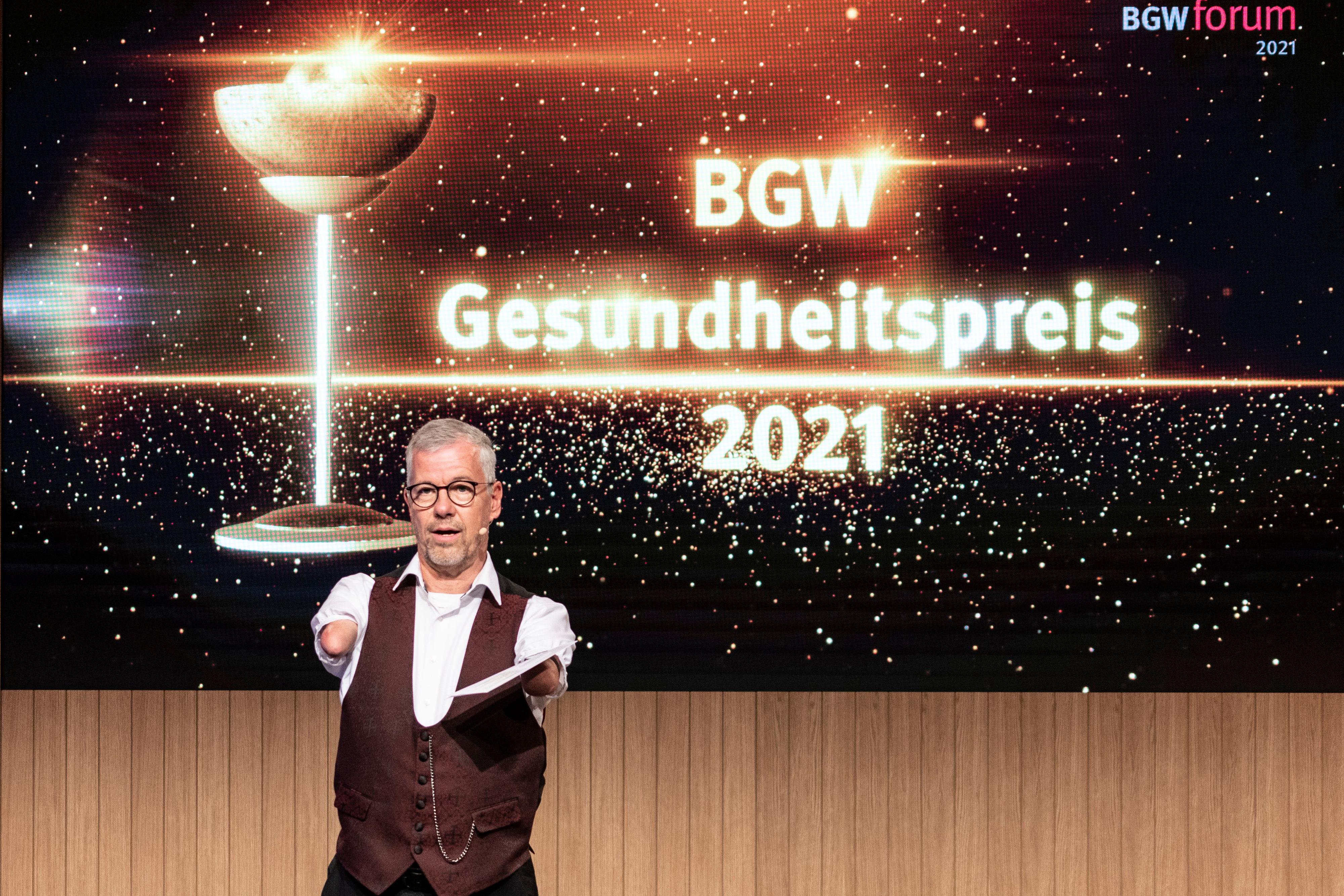  Moderator Rainer Schmidt vor einer Leinwand mit Text "BGW Gesundheitspreis 2021"