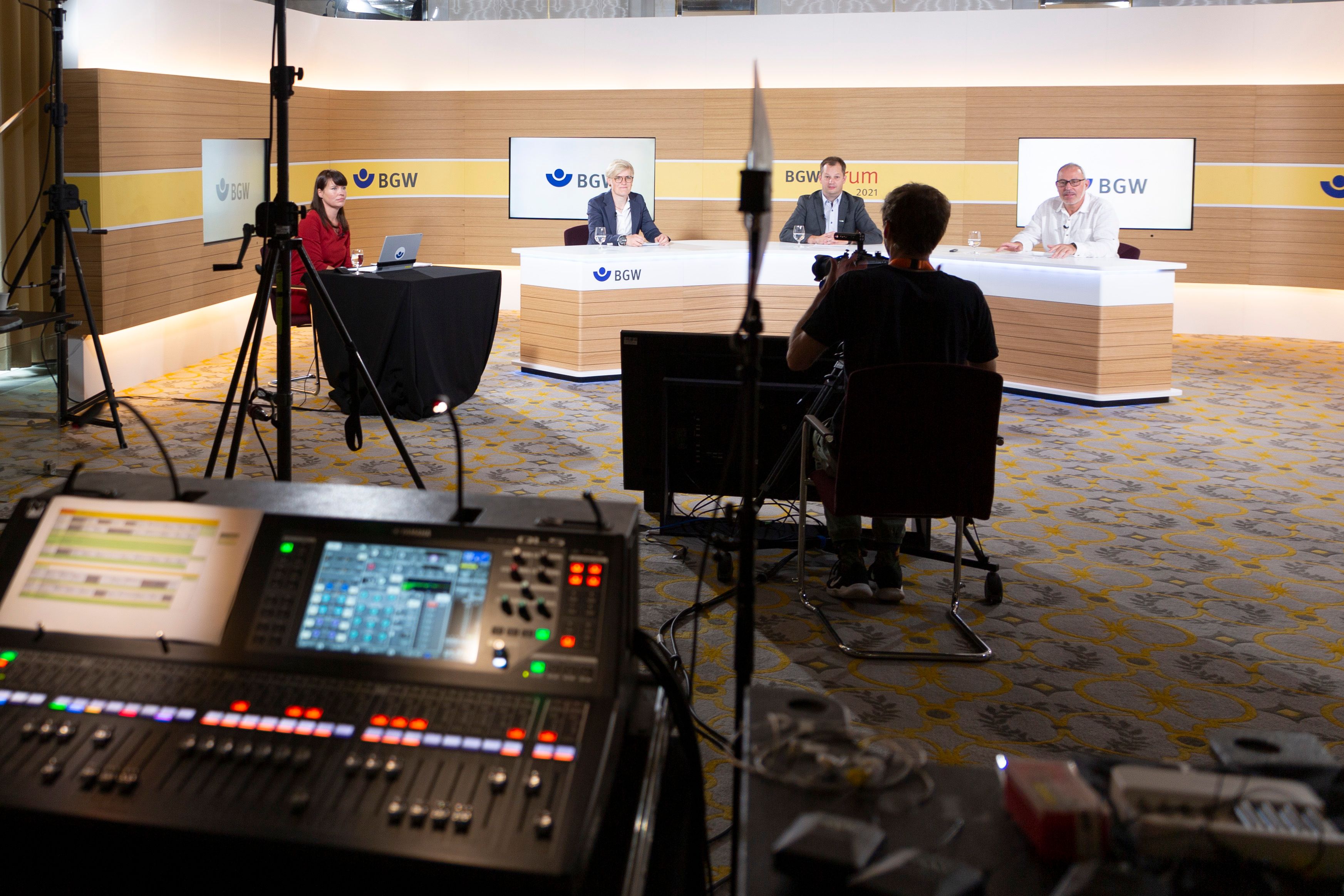 Studioansicht, im Vordergrund Monitore und Kameras, hinten sitzen Menschen an einem Tresen und werden gefilmt