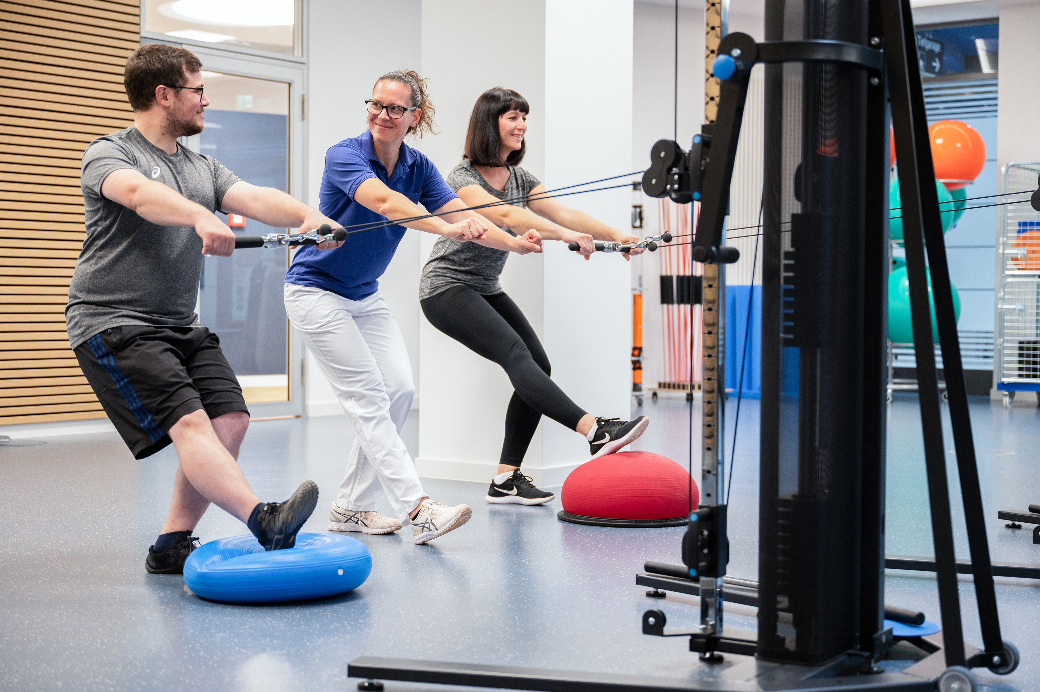 Ein Mann und eine Frau üben jeweils an einer Seilzugmaschine im Fitnessraum. Eine Therapeutin steht links neben dem Mann und korrigiert seine Haltung.