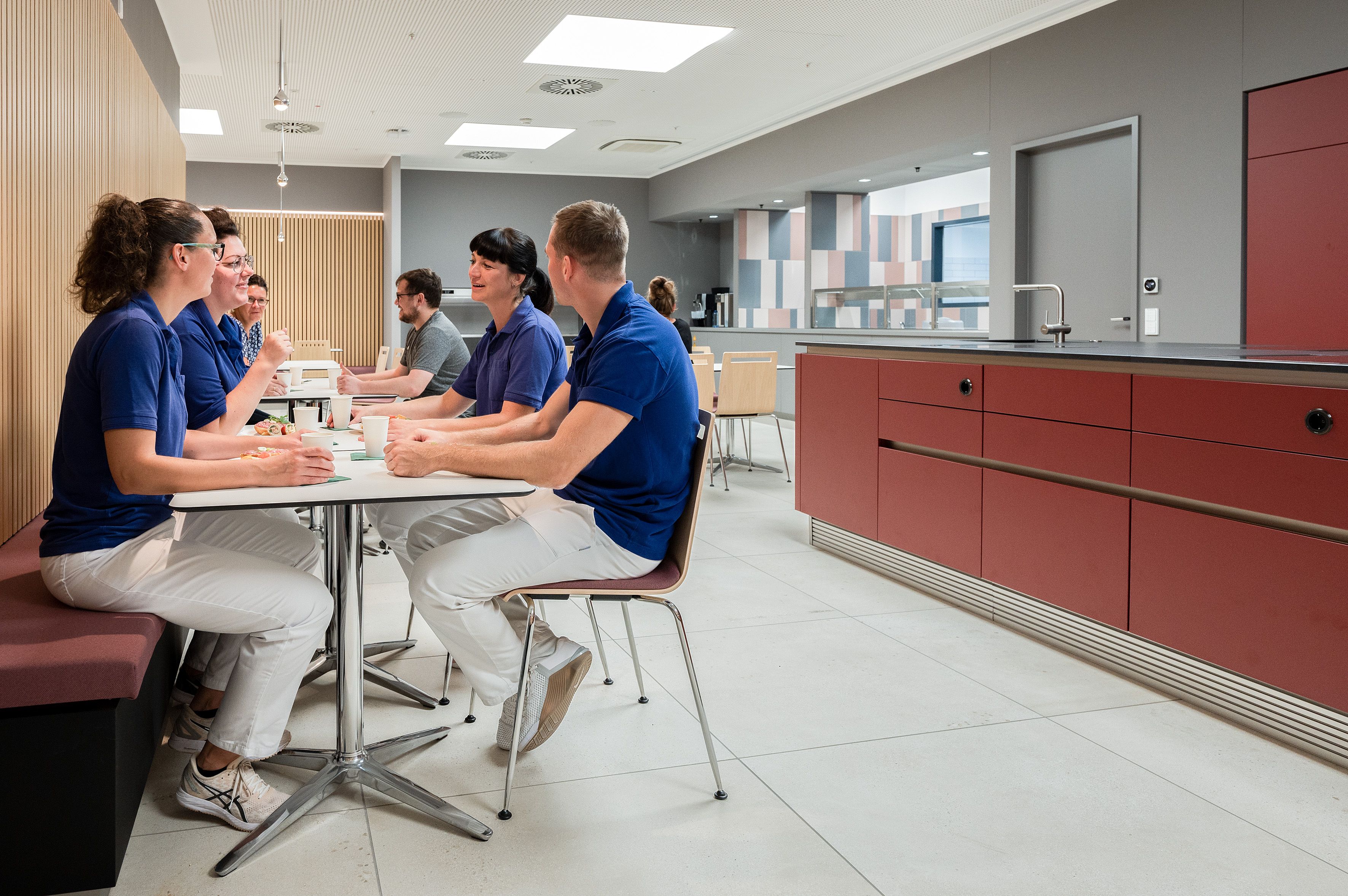 An einem Esstisch in einer Kantine unterhalten sich vier Personen. Sie tragen alle weiße Hosen und blaue Oberteile. Rechts steht eine dunkelrote Küchenzeile mit Küchentresen.