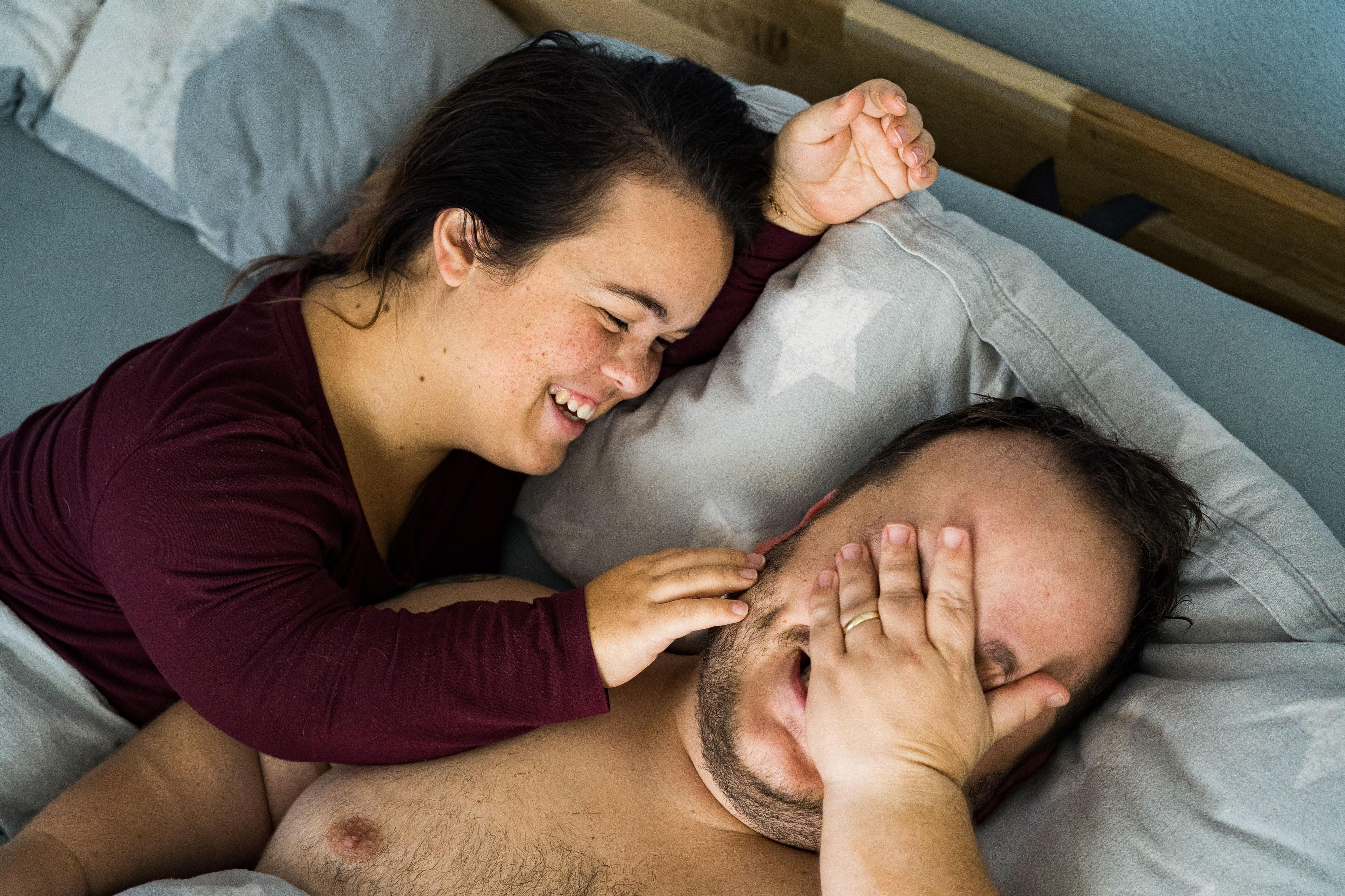 Ein junges Paar liegt nebeneinander im Bett. Sie streicht im zärtlich über die Wange. Er hat eine Hand vor dem Gesicht. Beide lachen.