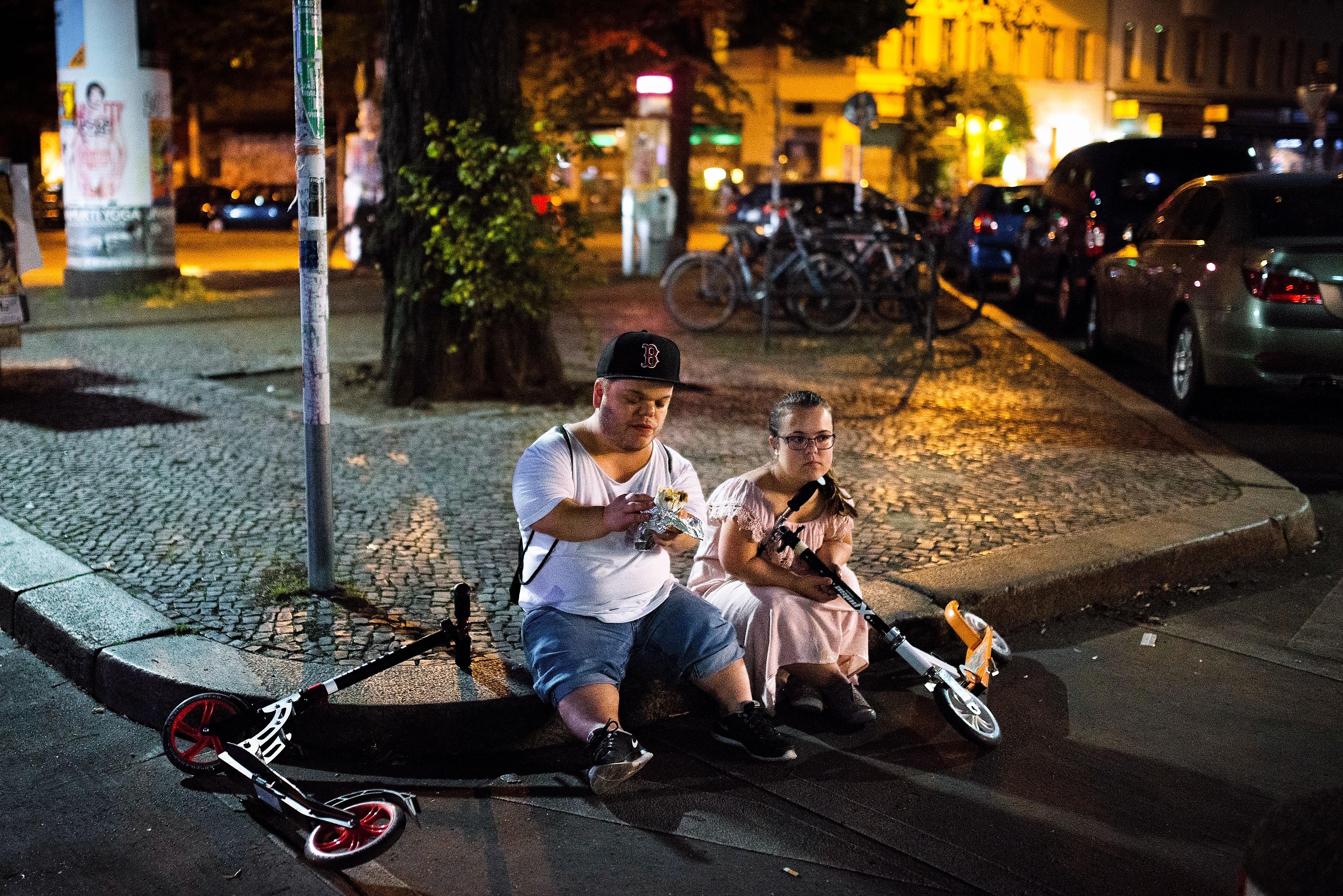 Ein kleinwüchsiges Paar sitzt auf der Bordsteinkante einer Stadt. Es ist dunkel. Neben ihnen liegen zwei Roller.