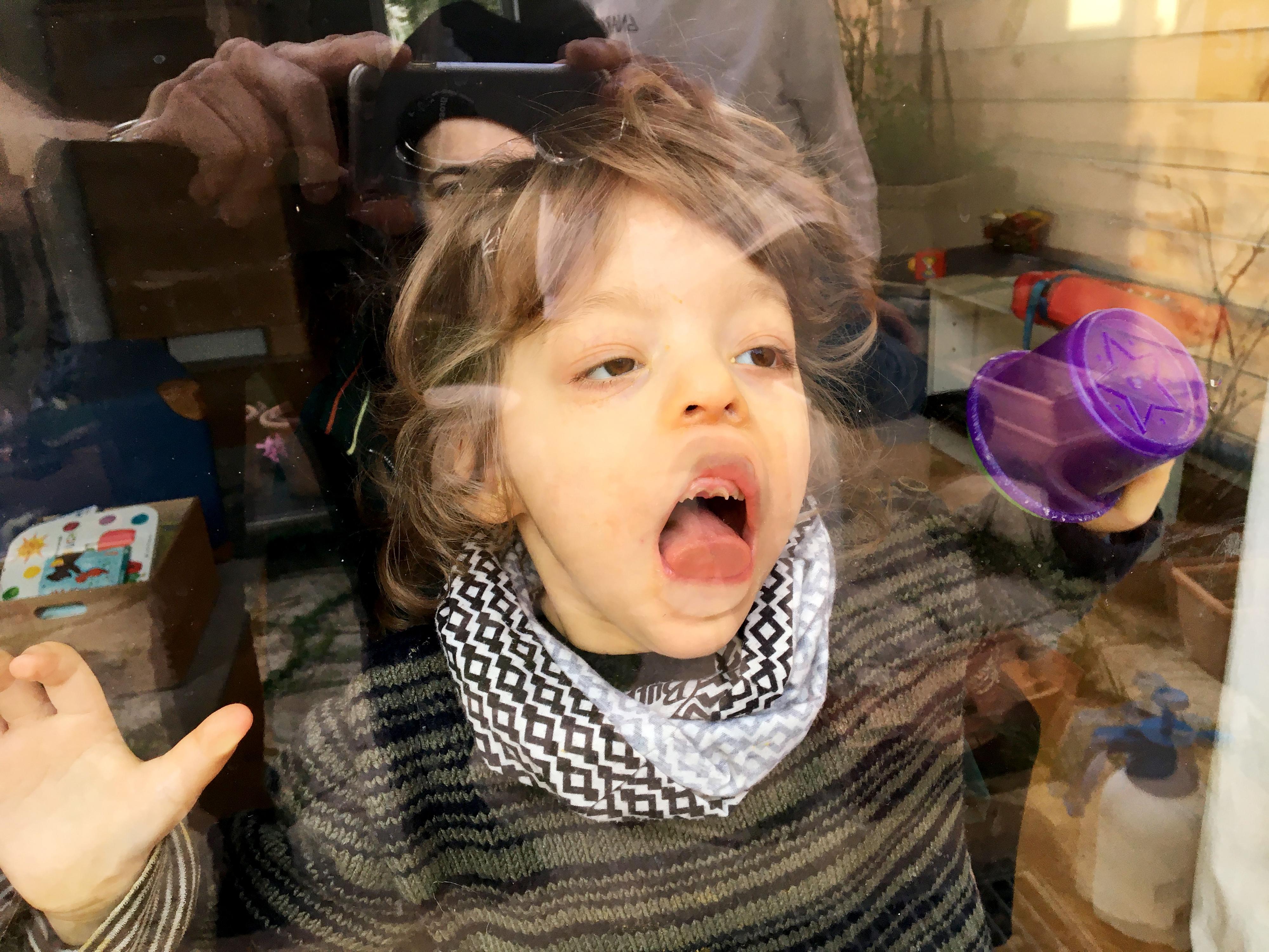 Mensch - Arbeit - Handicap 2021 Ein Kleinkind steht hinter einer Scheibe und leckt daran. Im Hintergrund stehen zwei andere Personen, die man nicht erkennt.