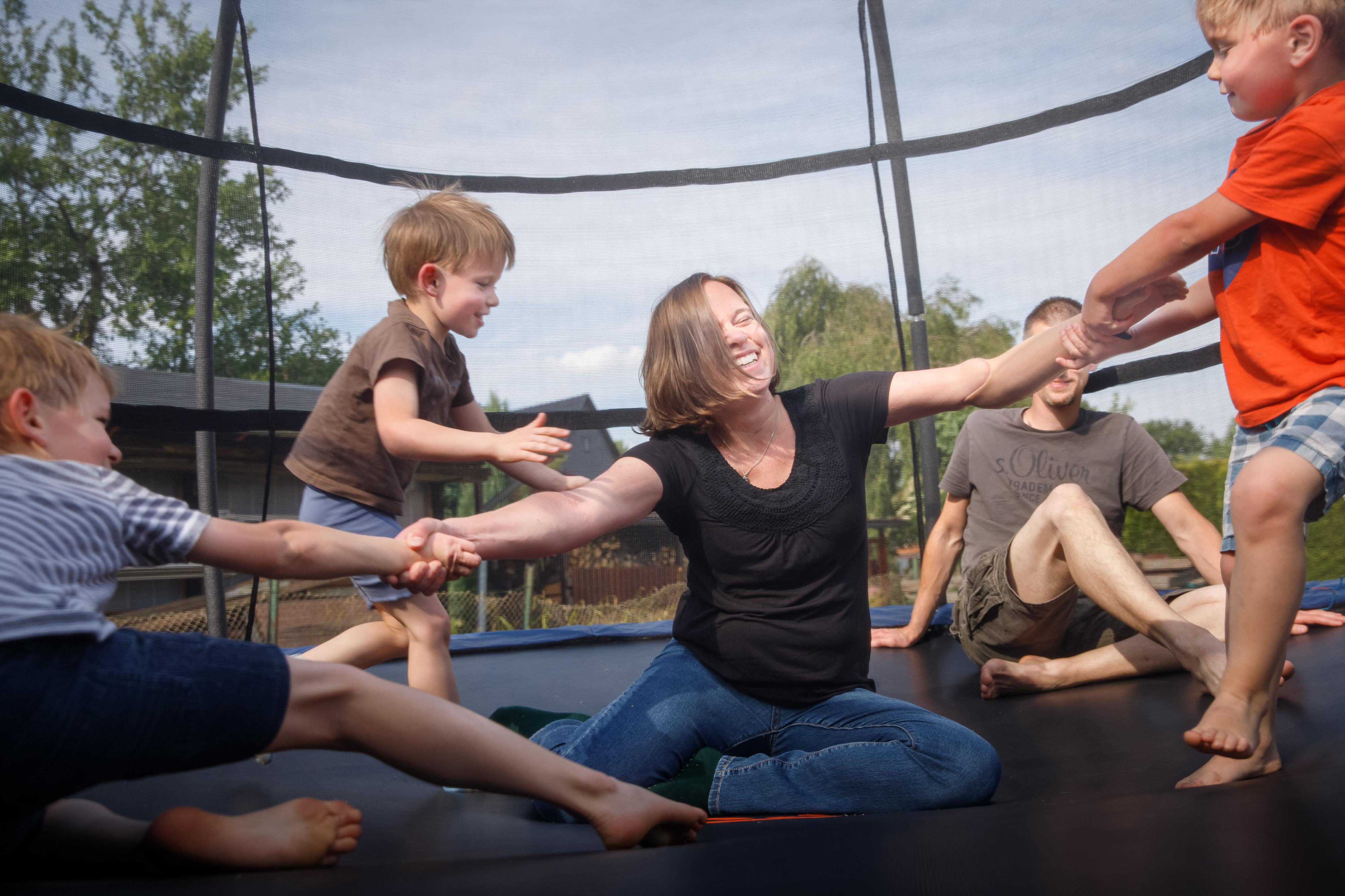 Eine Frau sitzt auf einem großen Trampolin. Um sie herum springen drei Kinder. Hinter ihr sitzt ein Mann.