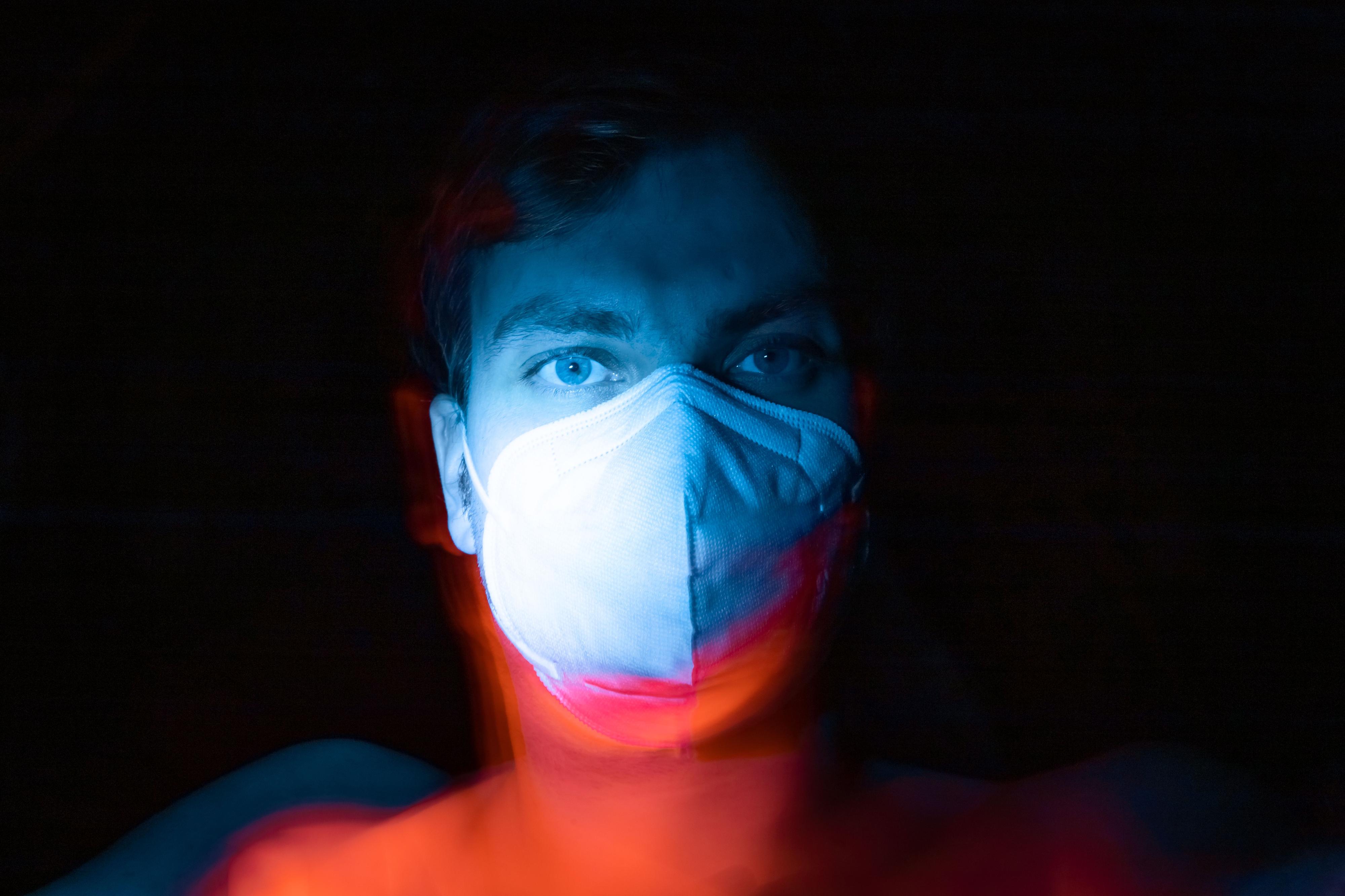 Ein Gesicht mit Mund-Nasen-Maske im Licht vor schwarzem Hintergrund.