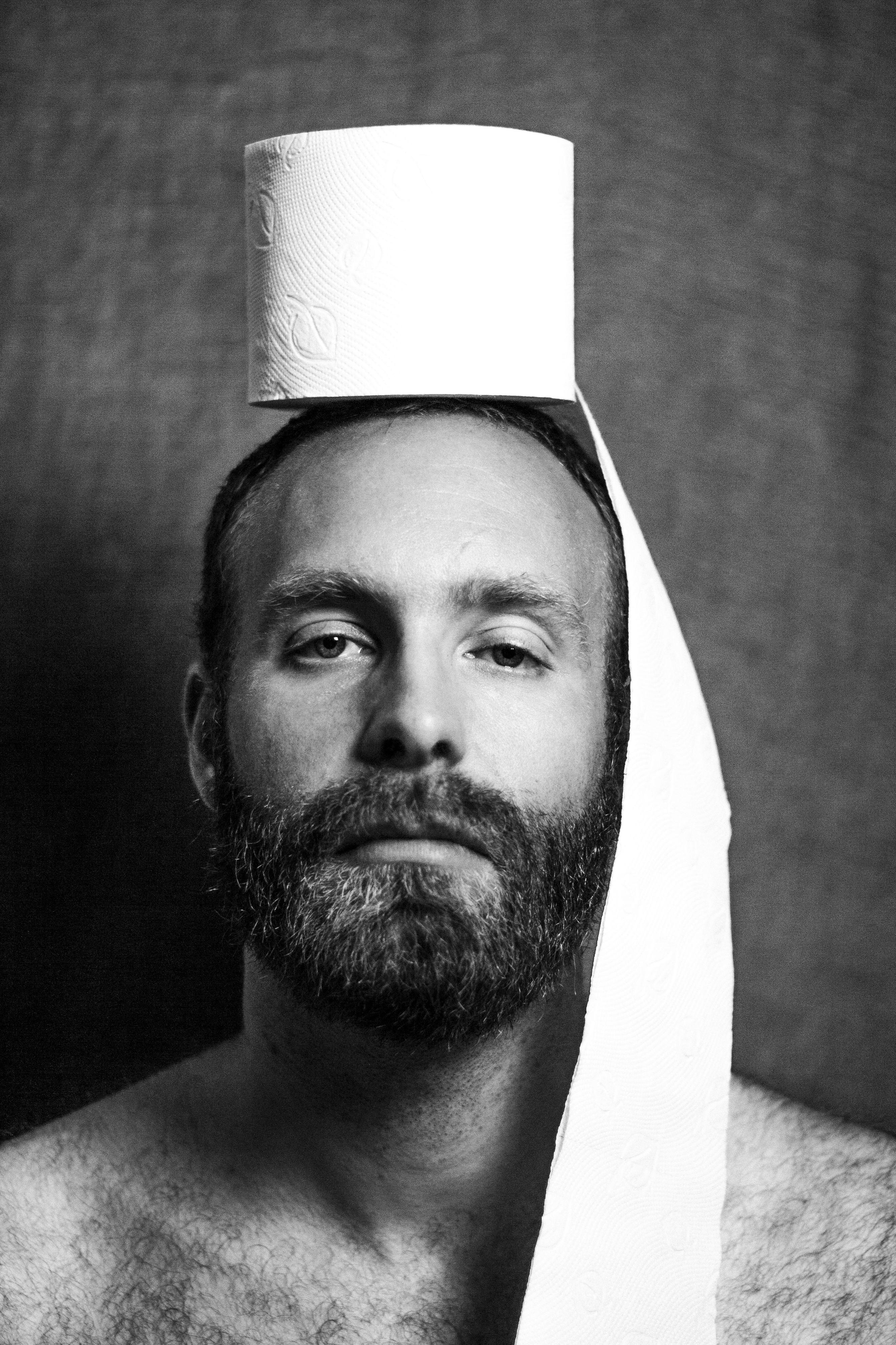 Porträt eines bärtigen Mannes. Auf seinem Kopf befindet sich eine Rolle Toilettenpapier - etwas davon fällt bis auf seine Schulter.