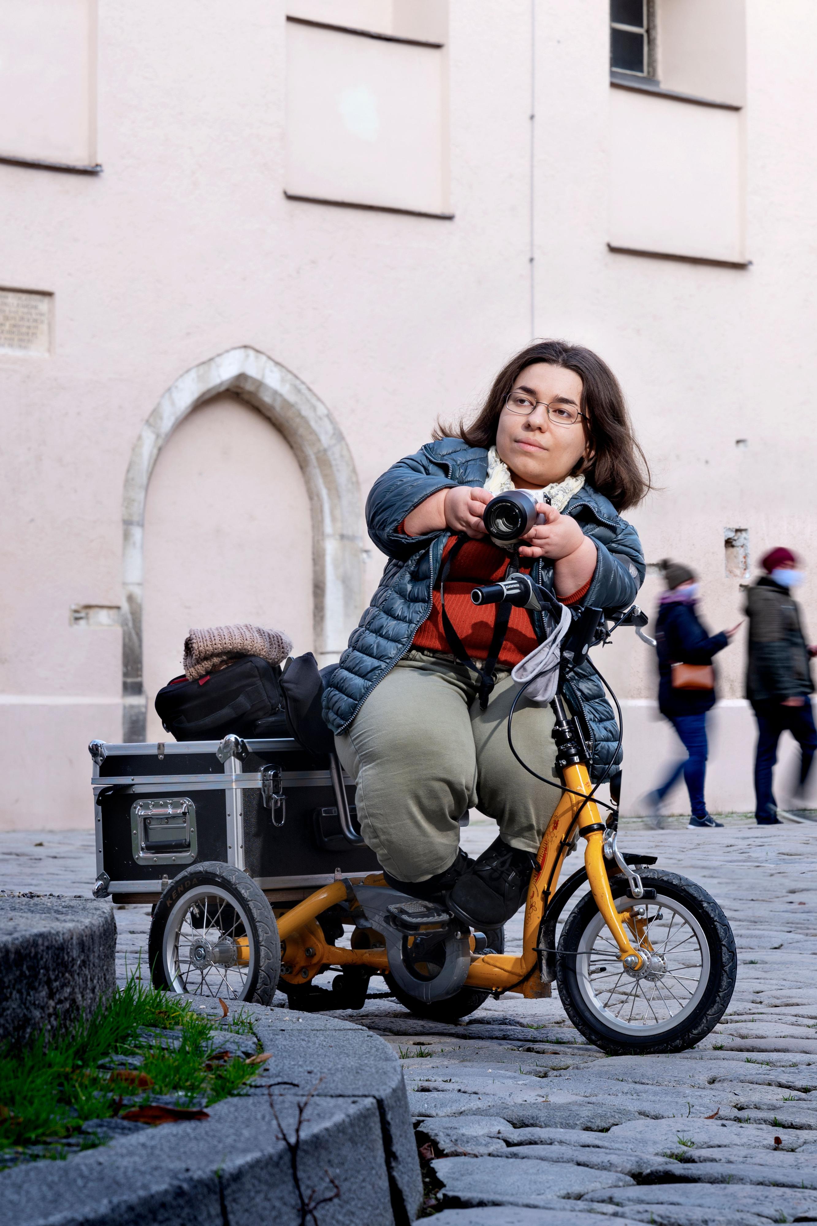 Eine kleinwüchsige Frau auf einem Roller steht auf einem Platz in der Stadt. Sie hält eine Kamera in der Hand.