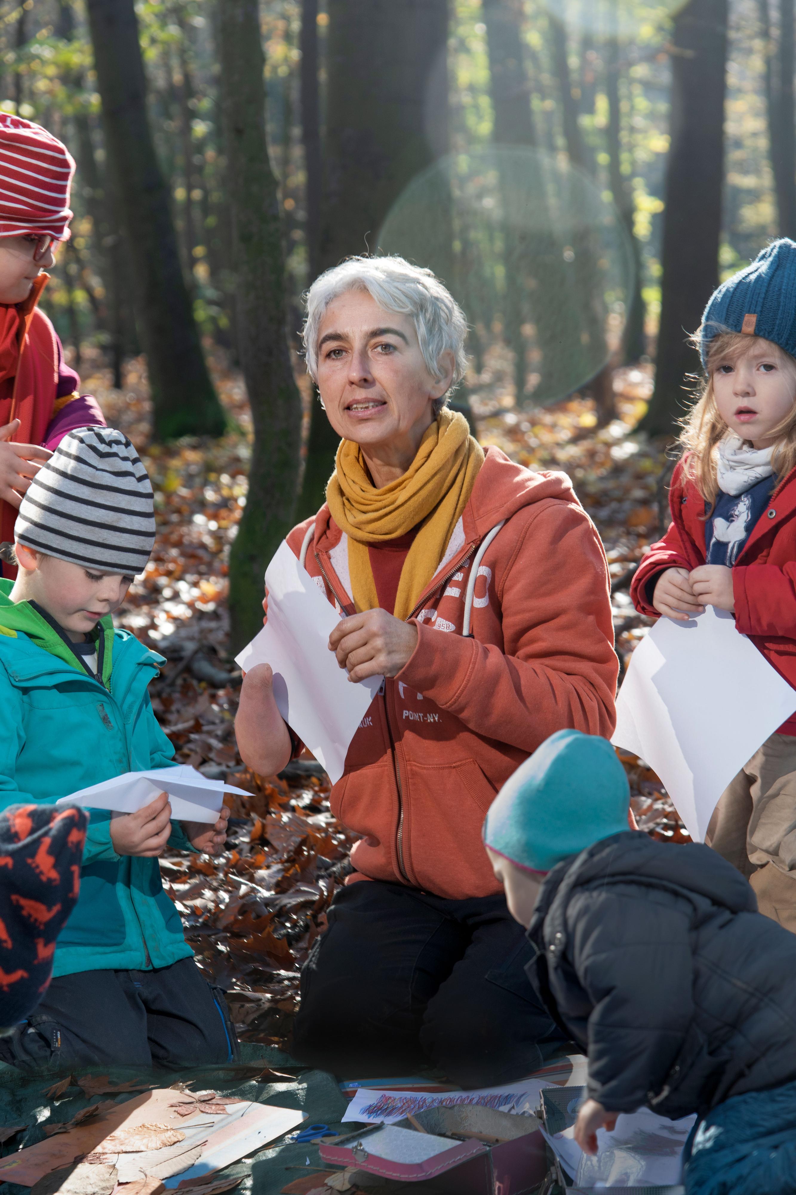 Eine Frau mit grauen Haaren sitzt mit vier Kindern im Wald. Sie bauen Papierflieger.