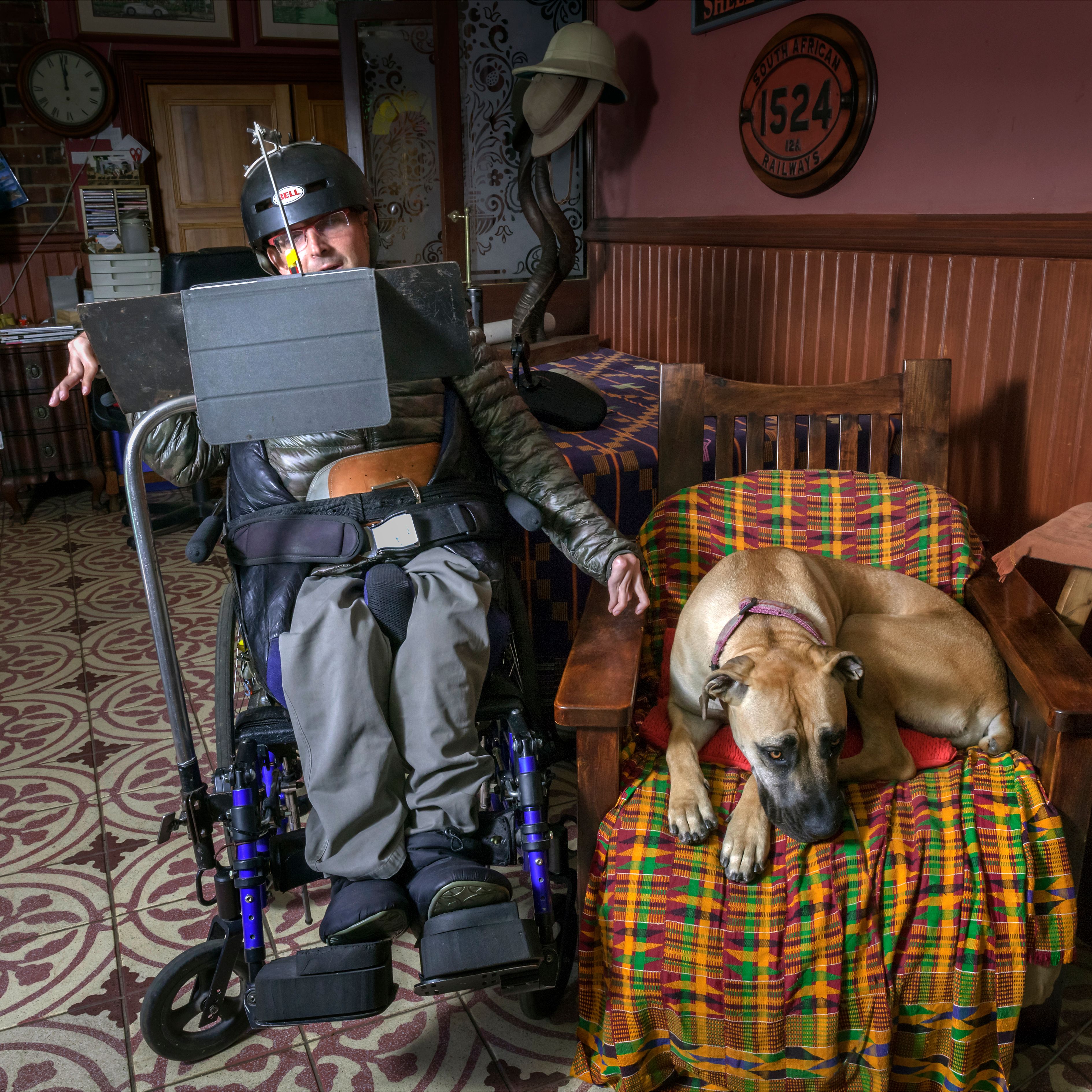 Mensch - Arbeit - Handicap 2021 Ein spastisch gelähmter Mann sitzt im Rollstuhl. Auf dem Kopf hat er einen Helm und vor sich einen Bildschirm. Daneben im Sessel liegt ein großer brauner Hund.