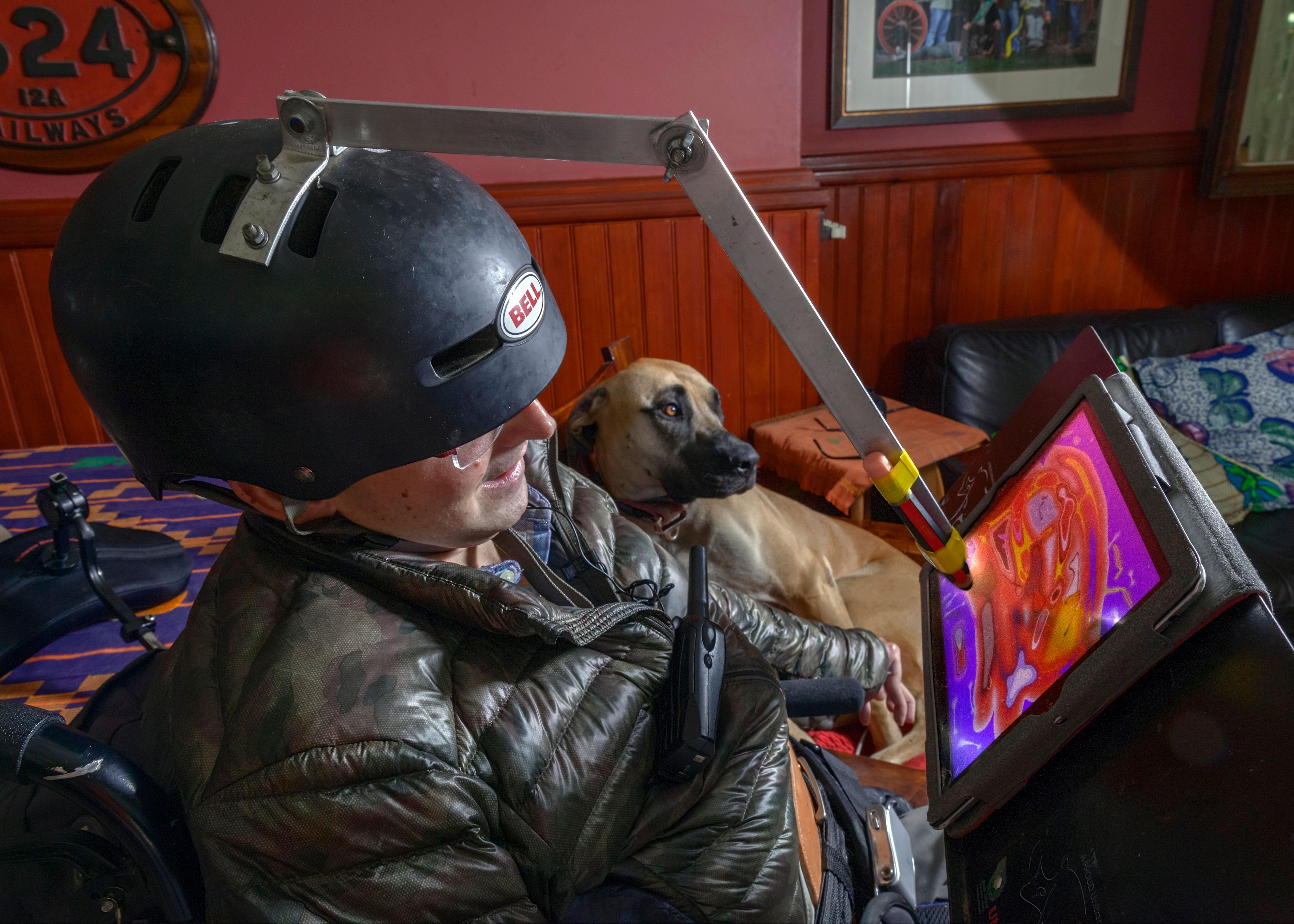 Ein Mann mit Helm auf dem Kopf. Der Helm hat eine Vorrichtung mit einem Stift, der Farben auf einen Bildschirm malt. Im Hintergrund liegt ein großer brauner Hund.