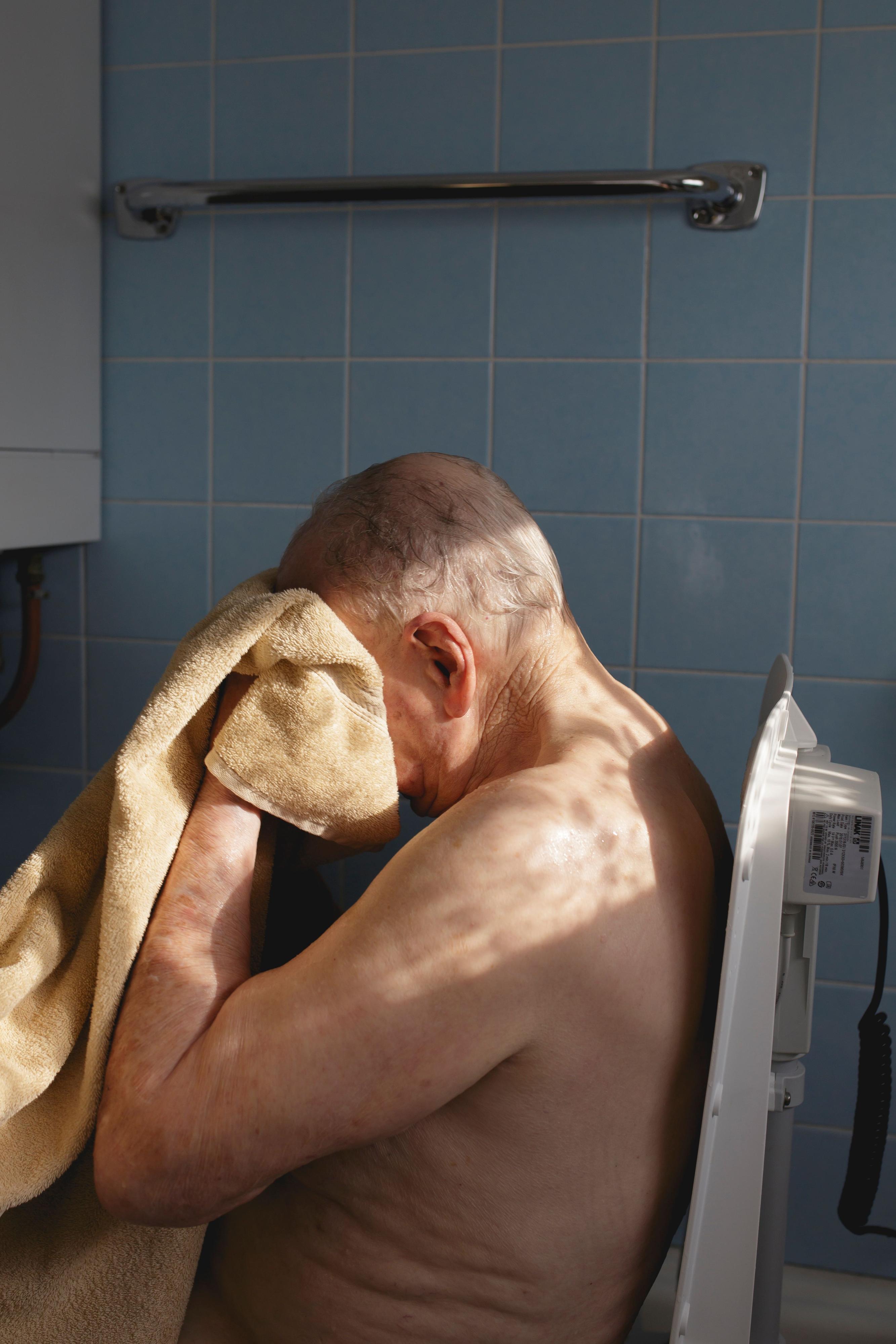 Ein älterer Mann sitzt mit nacktem Oberkörper in einem gekachelten Raum. Er trocknet sich mit einem Handtuch das Gesicht.