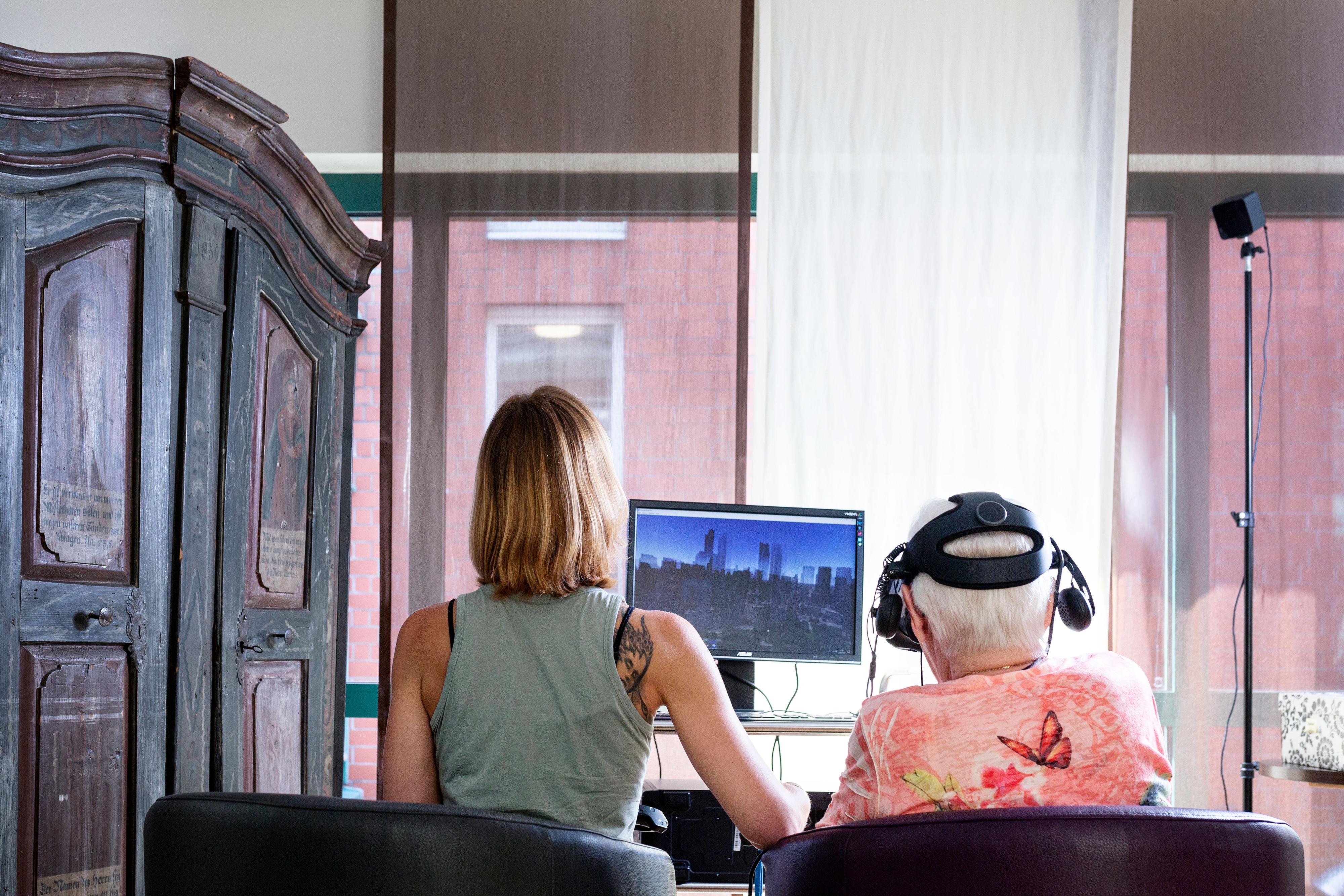 Eine ältere Frau mit VR-Brille sitzt neben einer jungen Frau vor einem Computer.