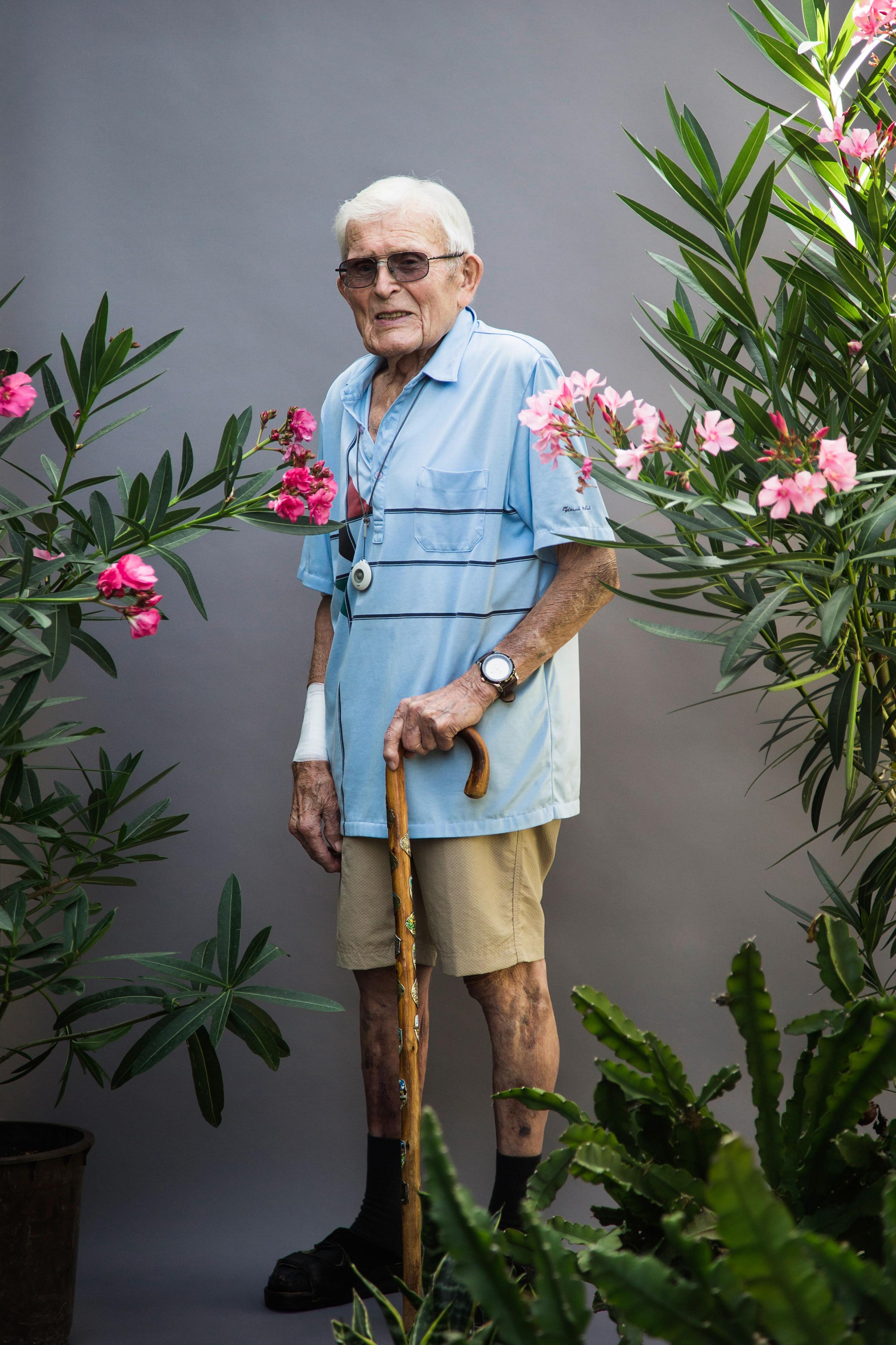 Ein älterer Mann steht mit Stock zwischen Pflanzen vor einer grauen Wand.