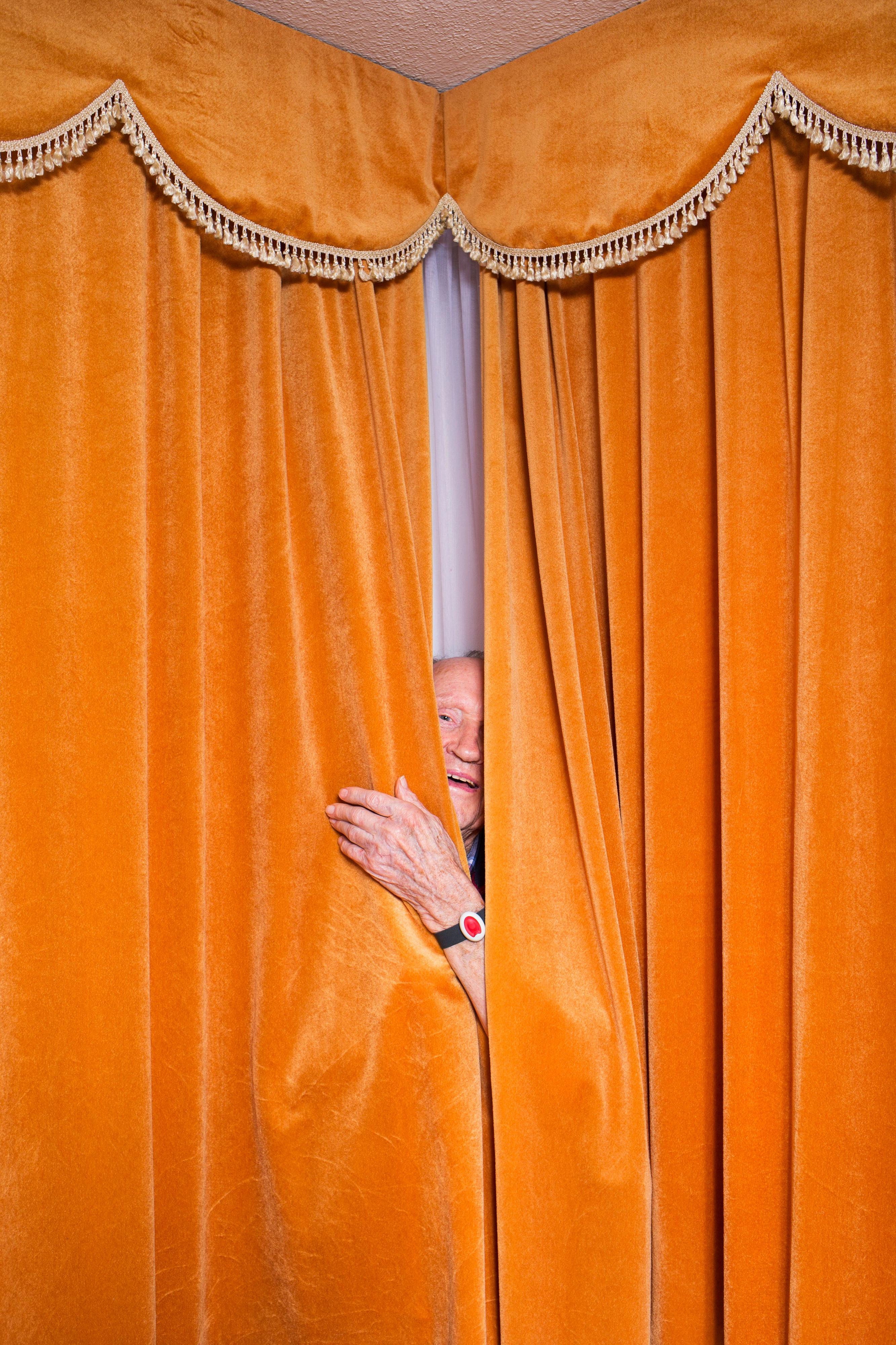 Ein älterer Mann schaut durch einen Schlitz in einem orangenen Vorhang.