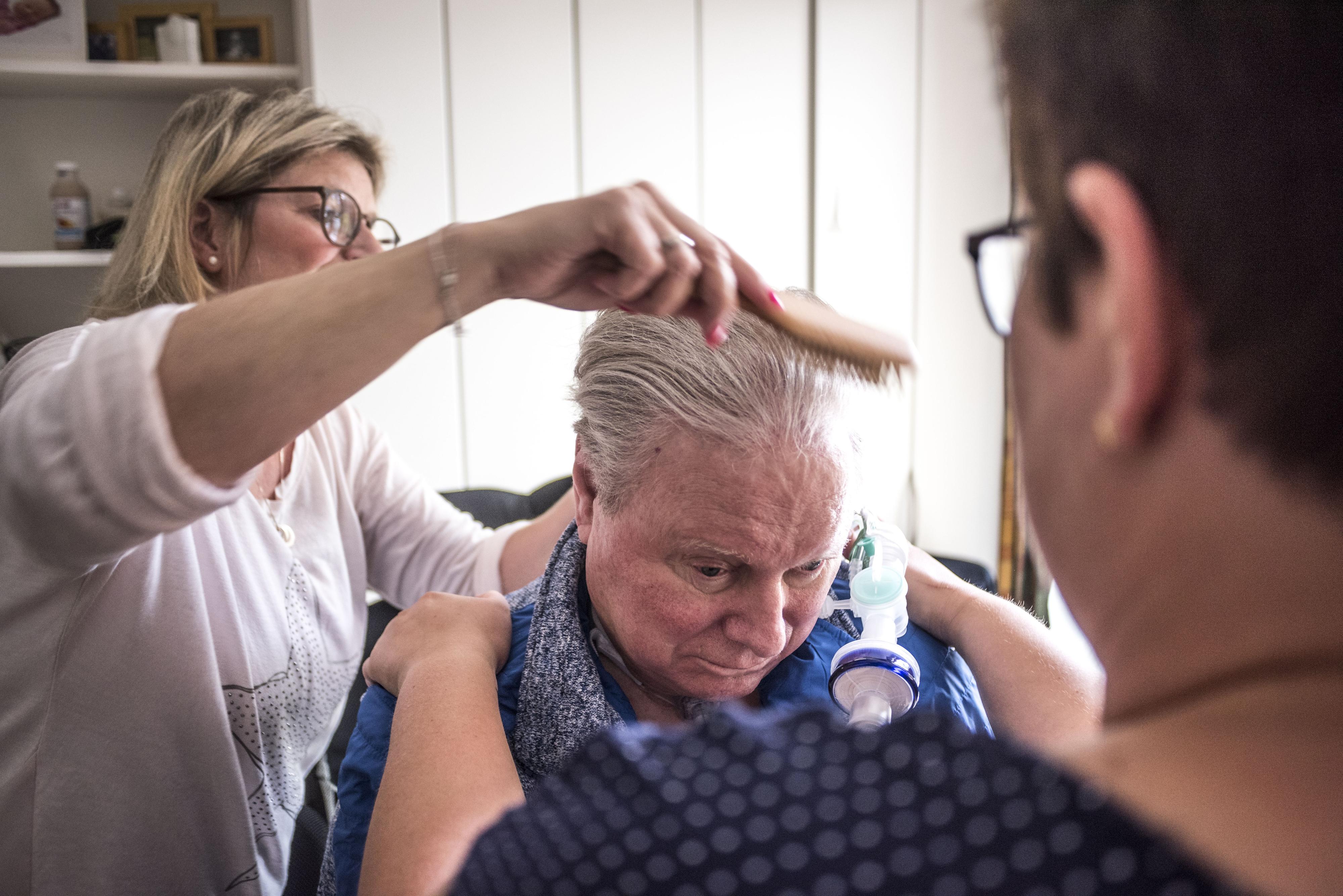 Eine Frau steht hinter einem älteren Mann und kämmt seine Haare. Eine andere Frau steht vor ihm und hält ihn an den Schultern.