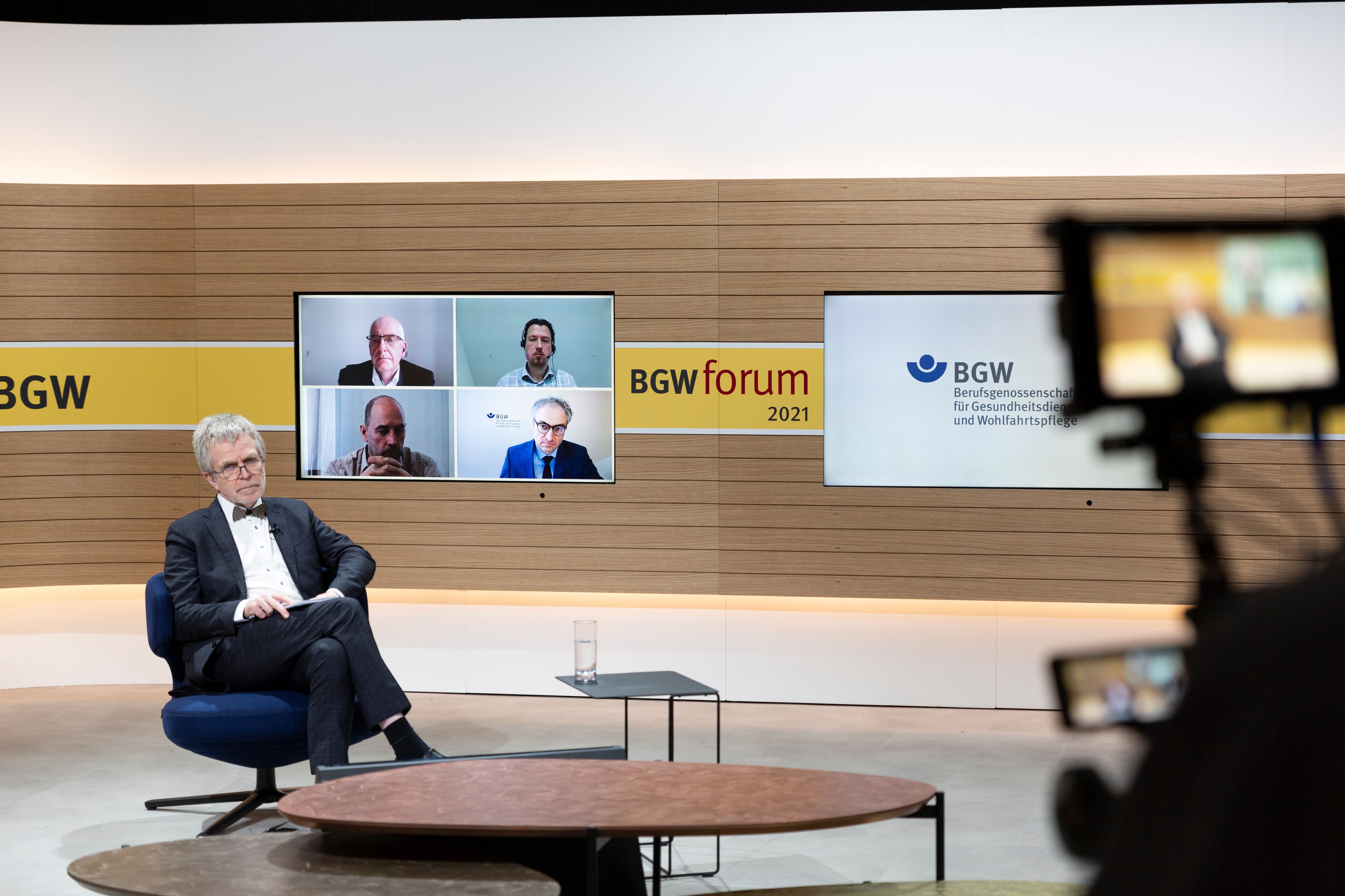 Screenshot: Der Moderator der Plenumsdiskussion sitzt in einem Studio - über einen Monitor an der Wand sieht man vier weitere Teilnehmer.