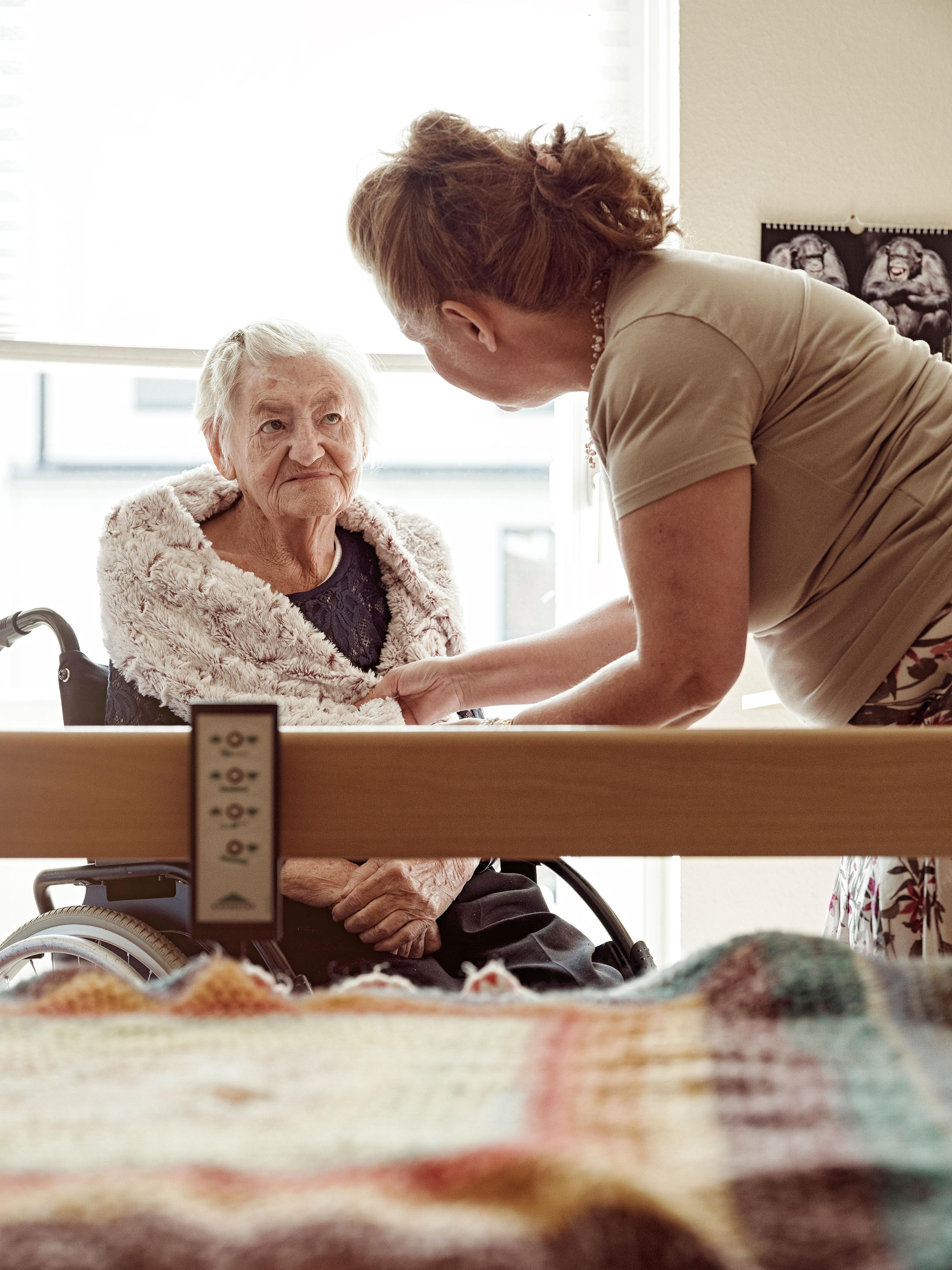 Eine ältere Frau sitzt im Rollstuhl vor einem Pflegebett und schaut einer jüngeren Frau in die Augen.
