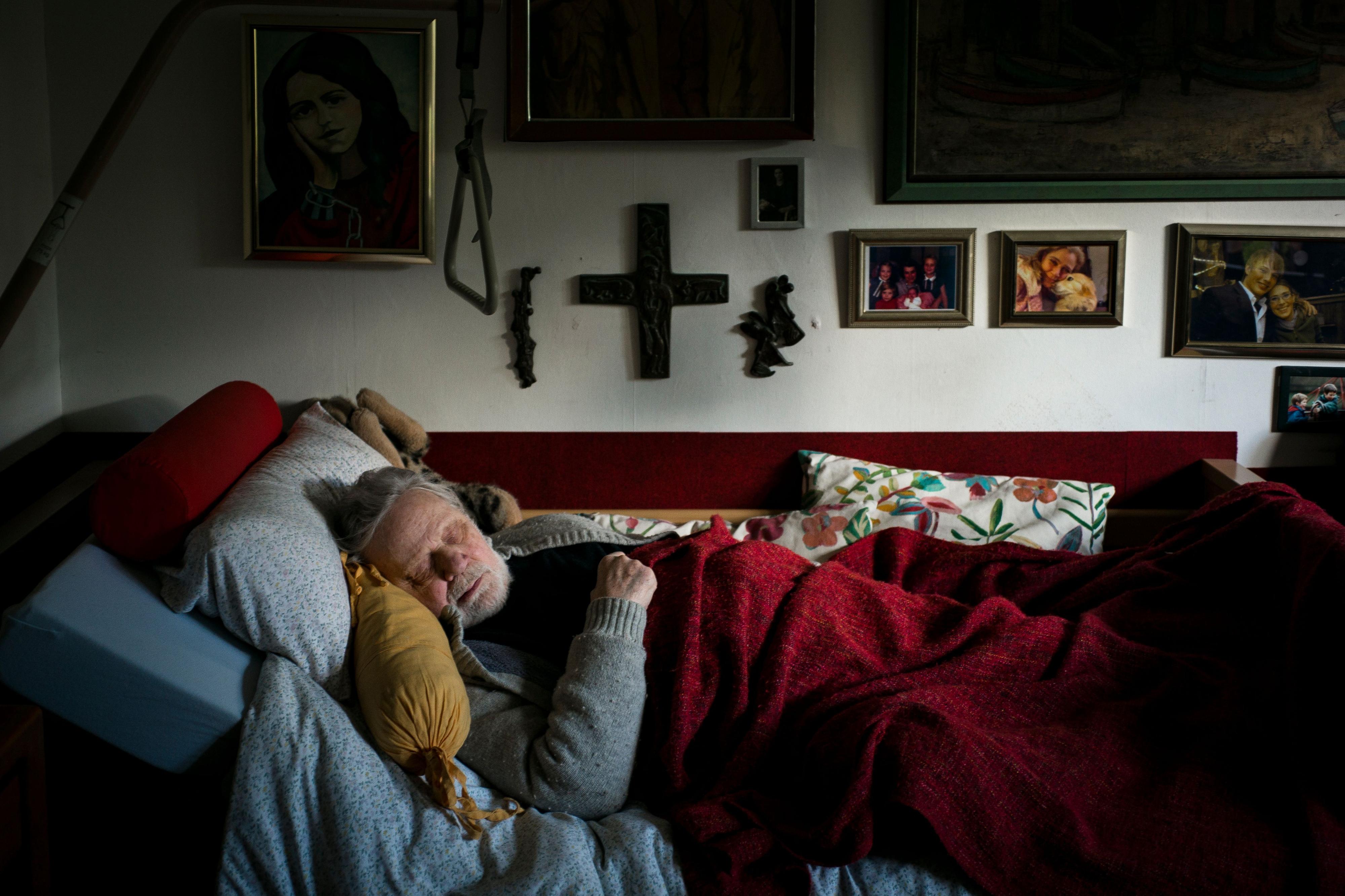 Ein Mann schläft in einem Bett vor einer Wand mit vielen Bilderrahmen.