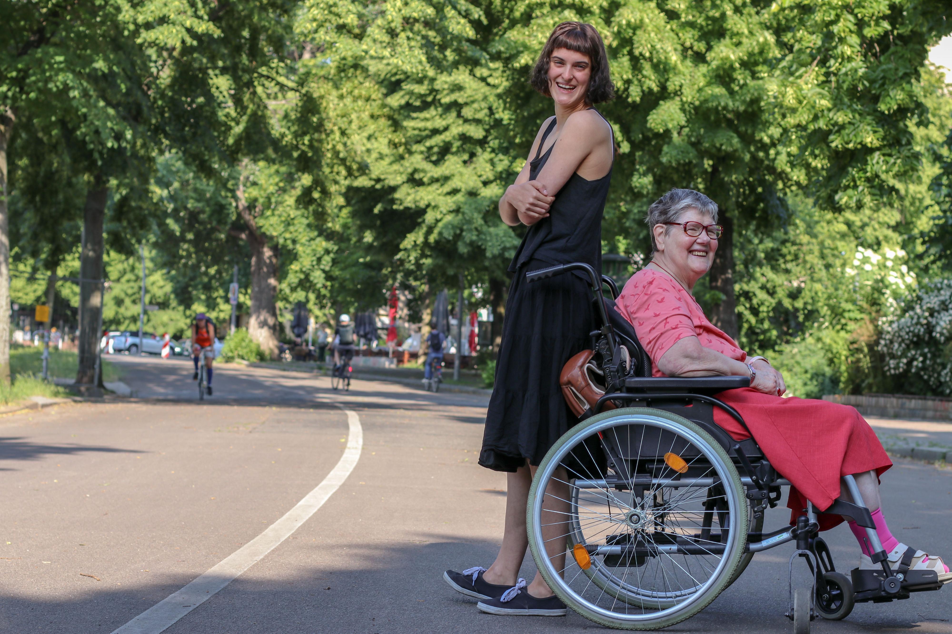 Eine junge Frau und eine ältere Frau im Rollstuhl stehen Rücken an Rücken auf einer Straße.