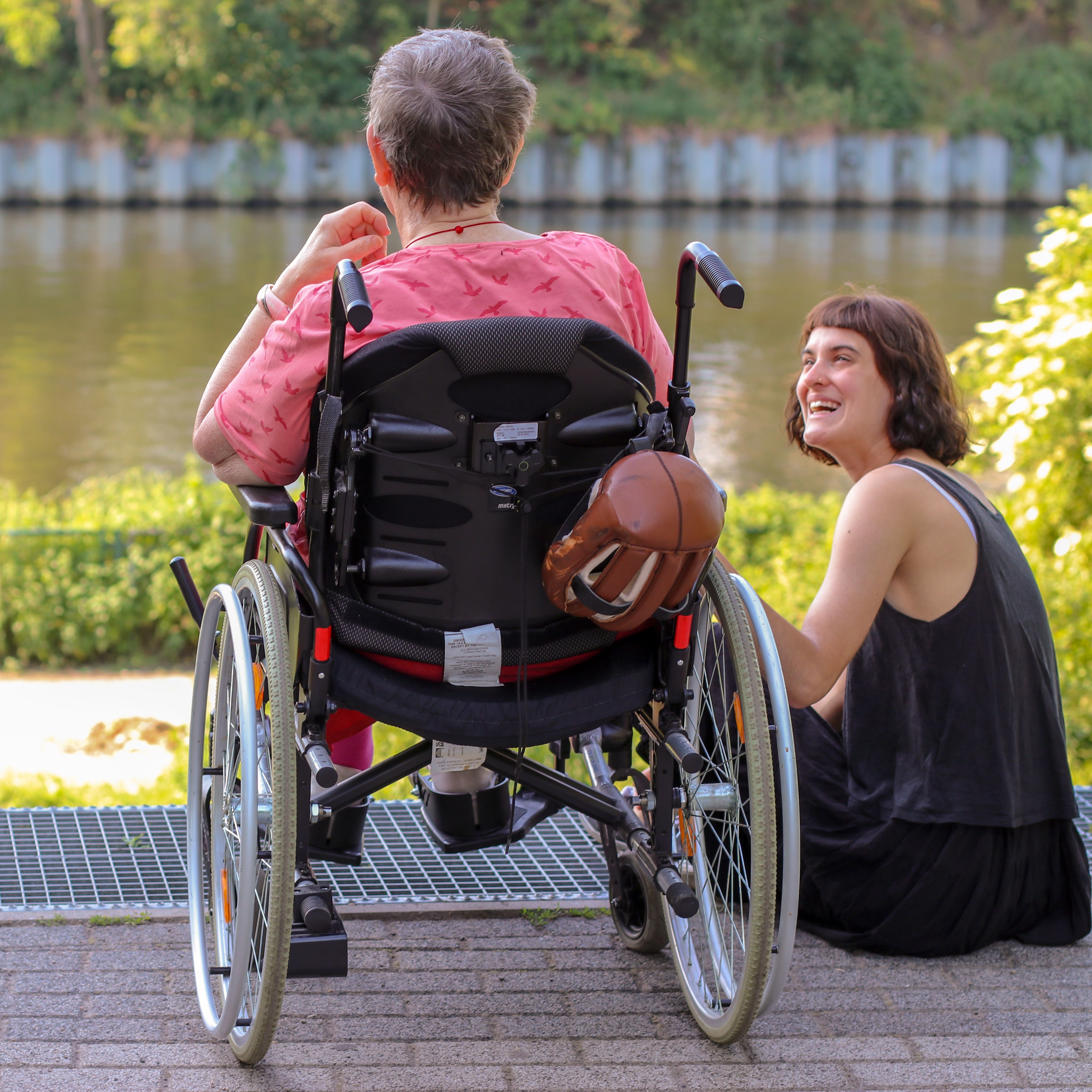 Eine alte Frau im Rollstuhl und eine junge Frau sitzen am Ufer eines Flusses.