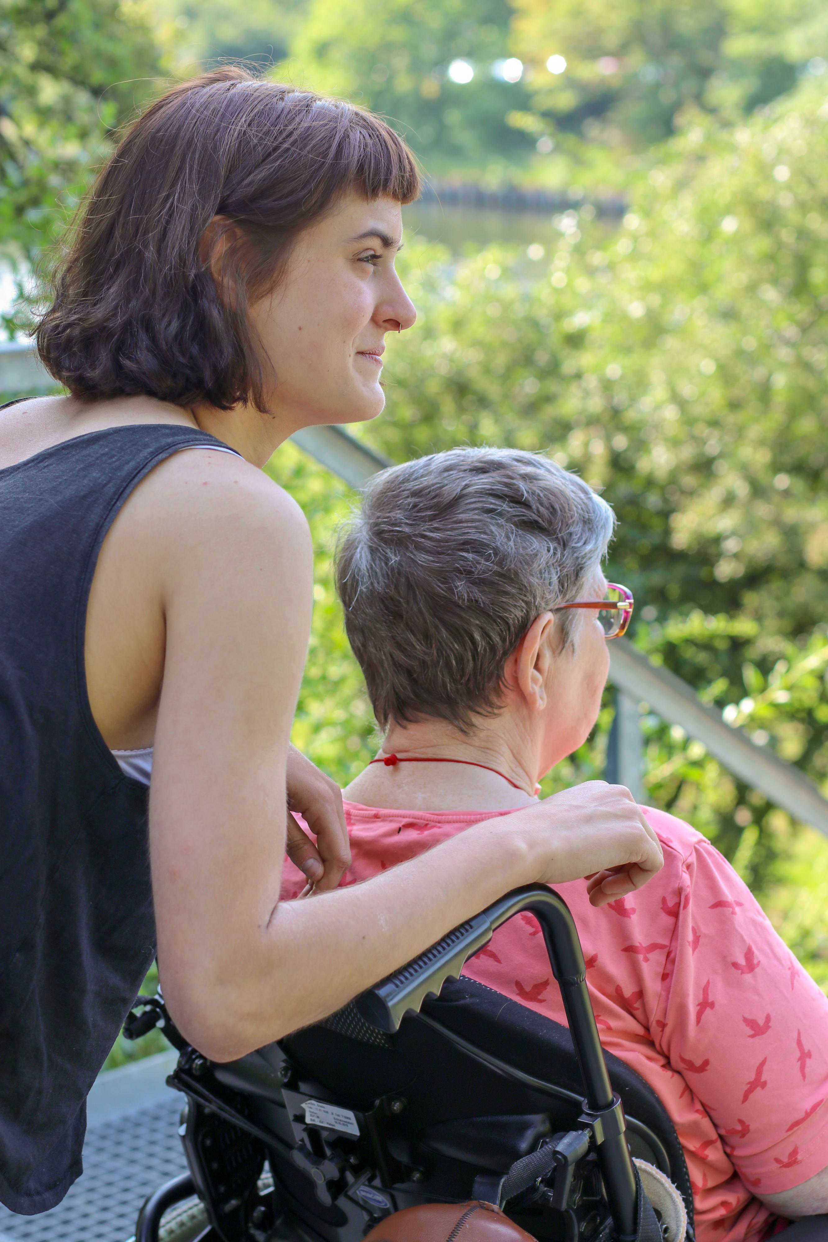 Eine ältere Frau sitzt im Rollstuhl, eine jüngere Frau lehnt sich lächelnd an.