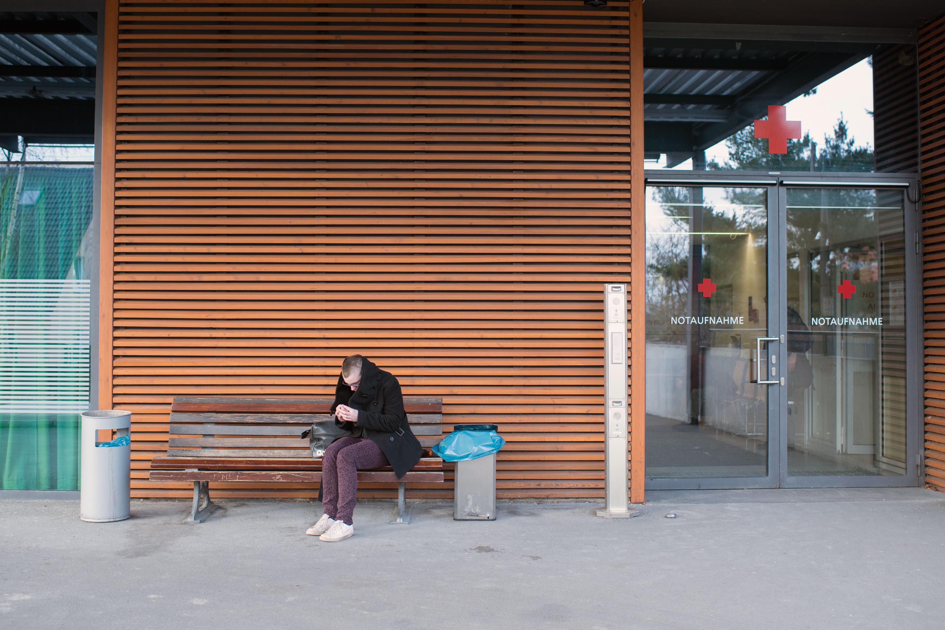 Eine junge Frau sitzt auf einer Bank vor einer Klinik.