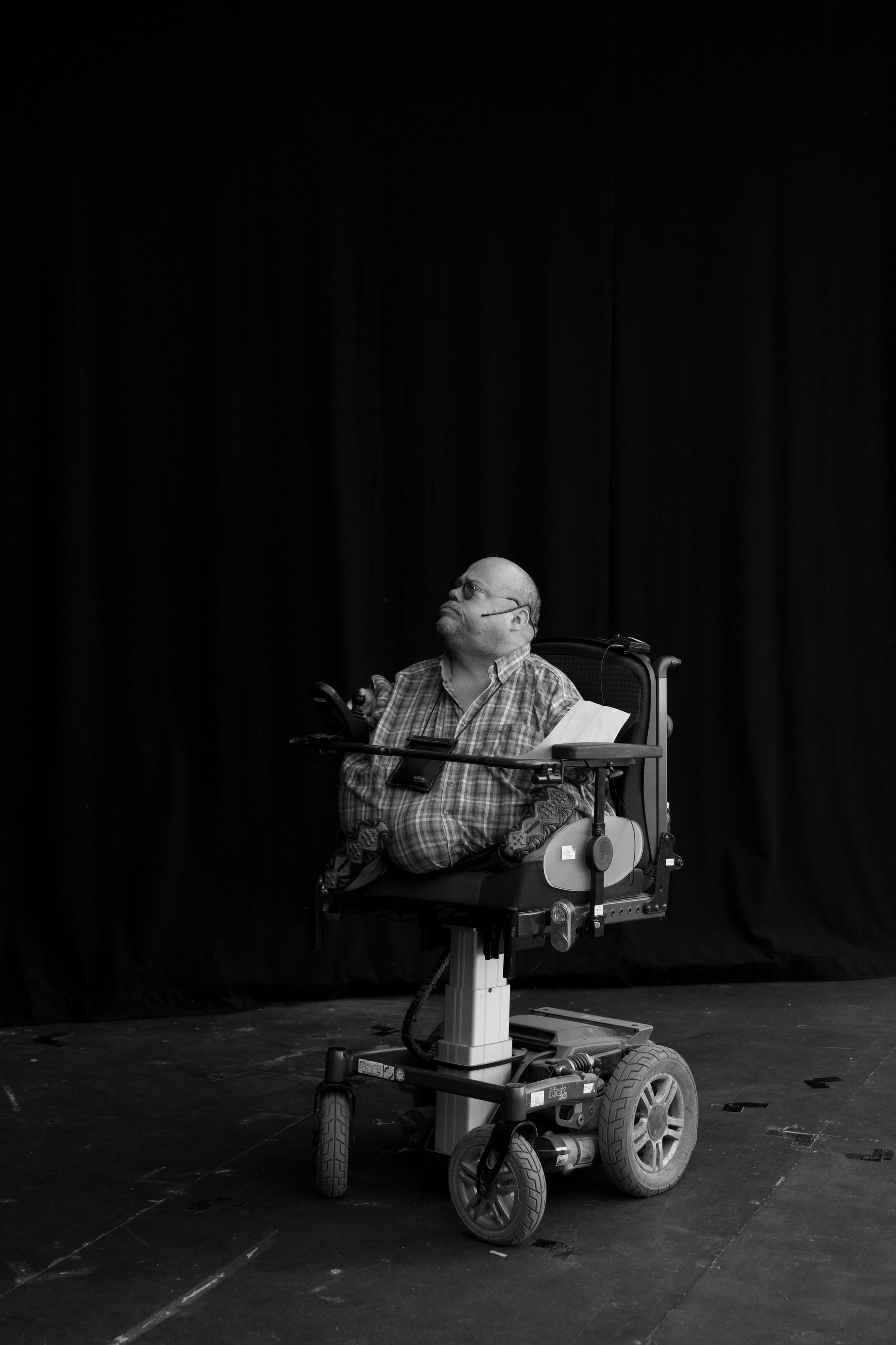 Ein Mann mit sehr kurz gewachsenen Armen und Beinen sitzt in einem Spezialrollstuhl auf einer Bühne.