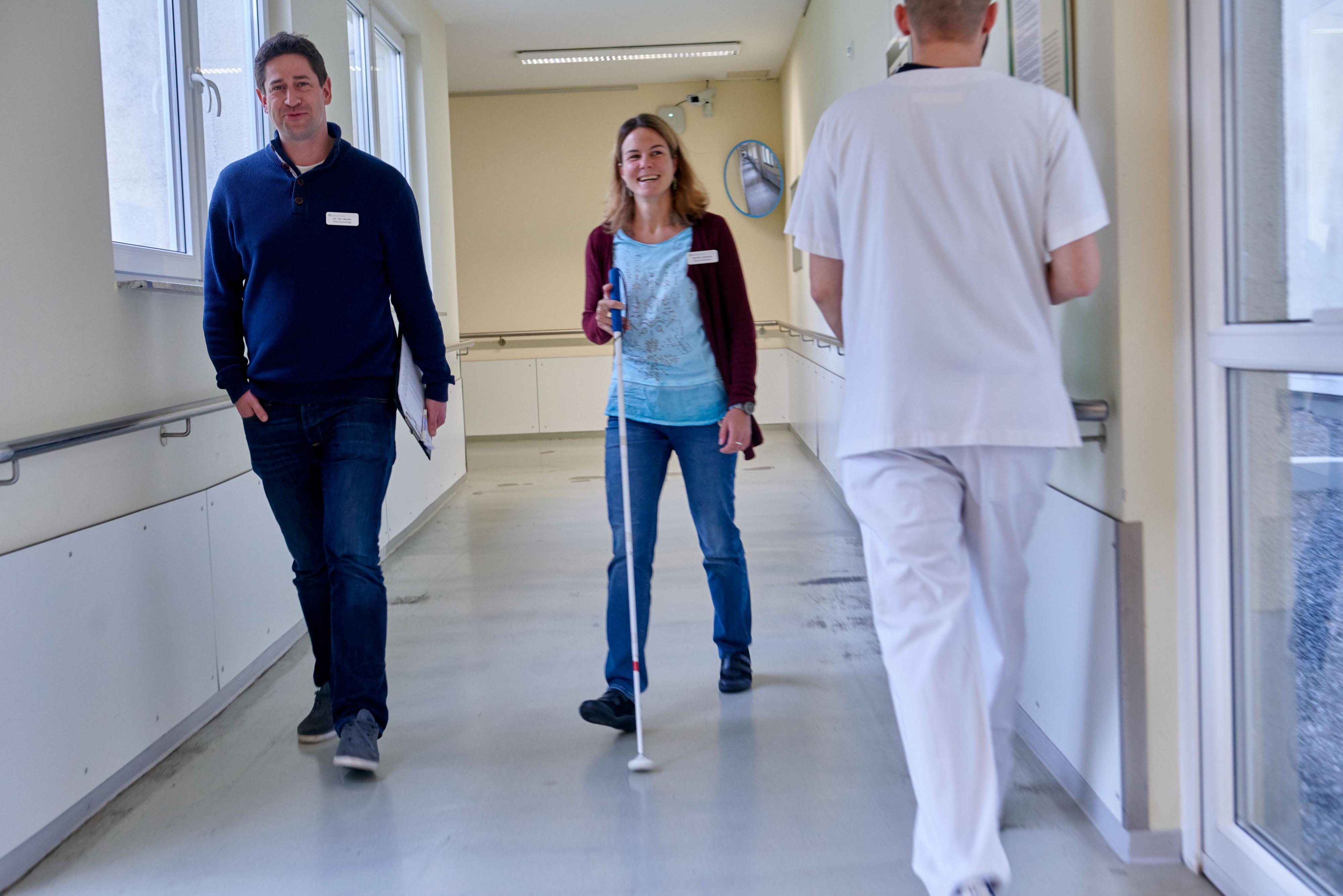 Ein Mann und eine Frau gehen einen Klinikflur entlang – die Frau trägt einen Blindenstock.