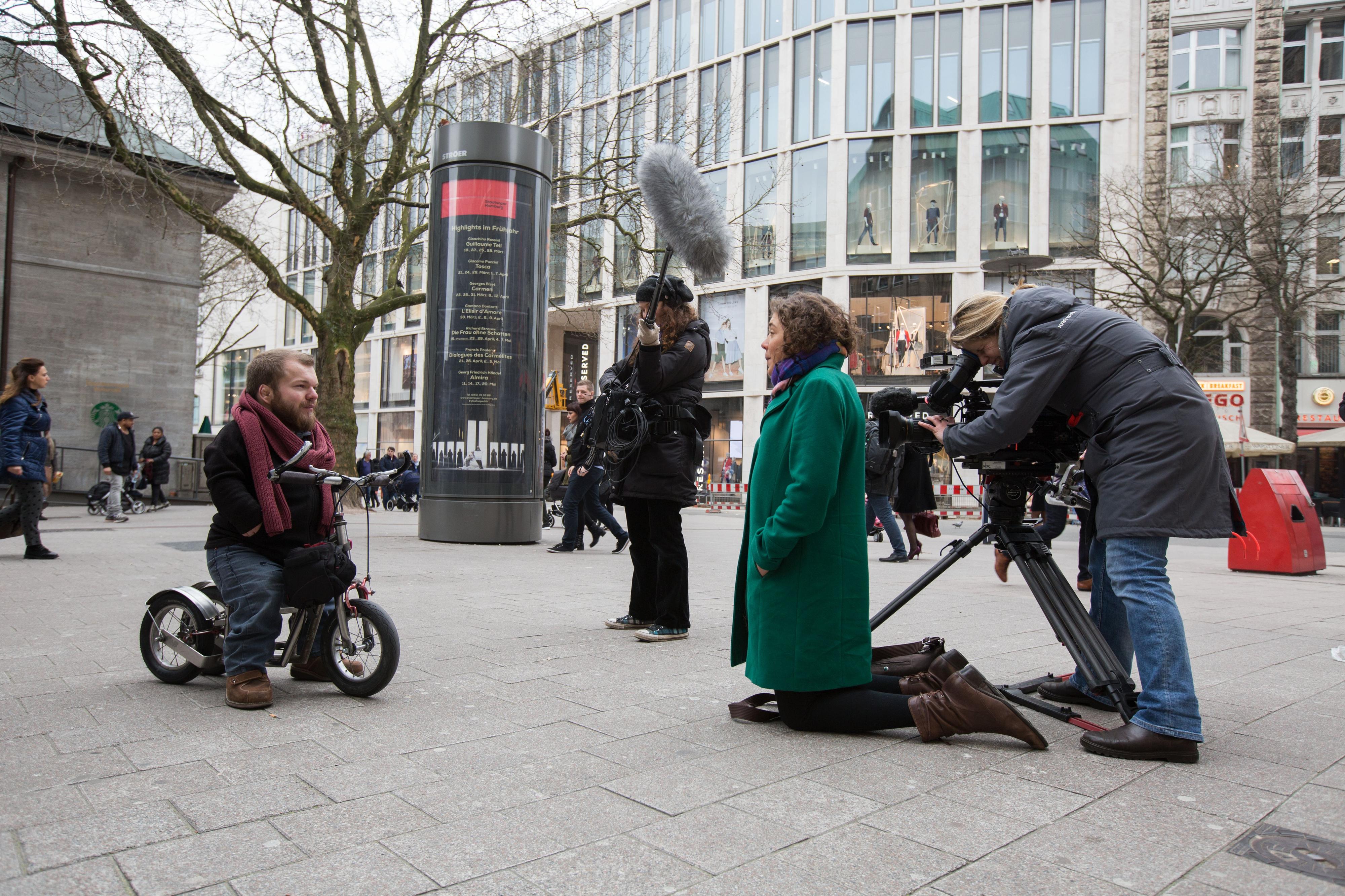 Ein kleinwüchsiger Mann sitzt auf einem Roller und wird von einem Fernsehteam in einer Einkaufsstraße gefilmt.