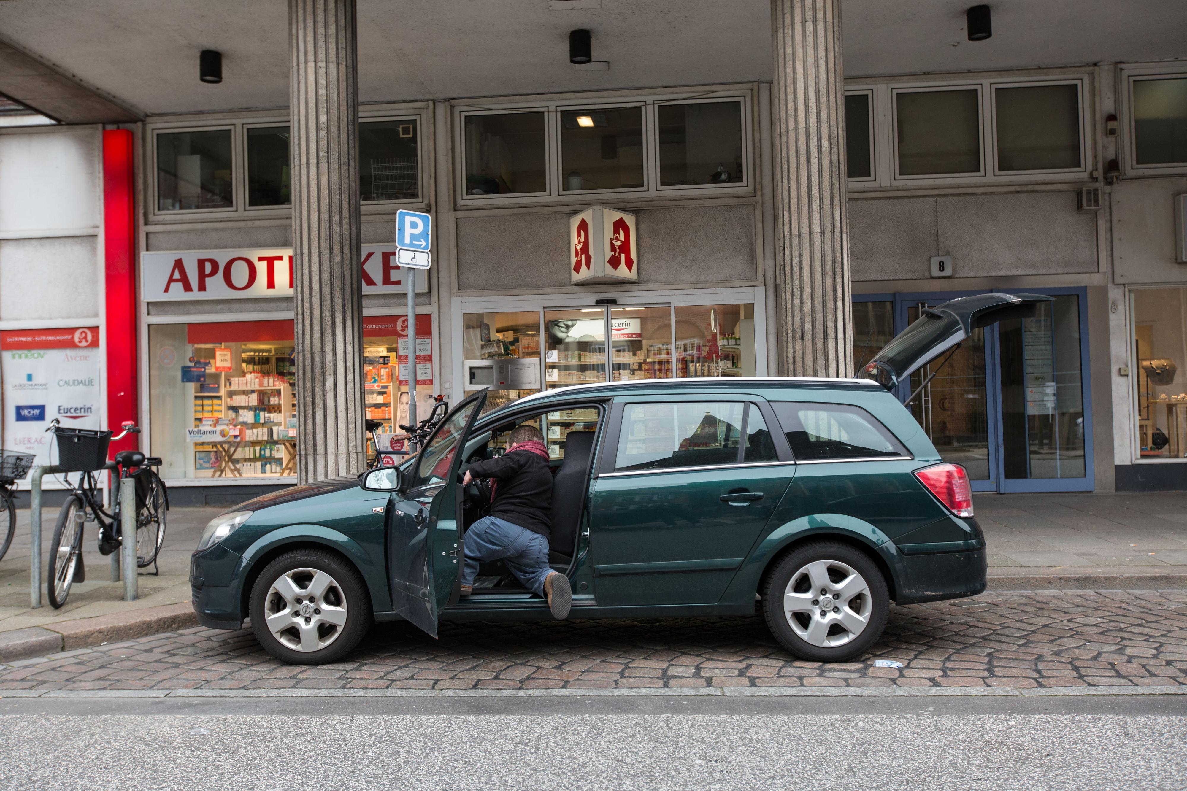 Ein kleinwüchsiger Mann steigt in ein Auto, das vor einer Apotheke steht.