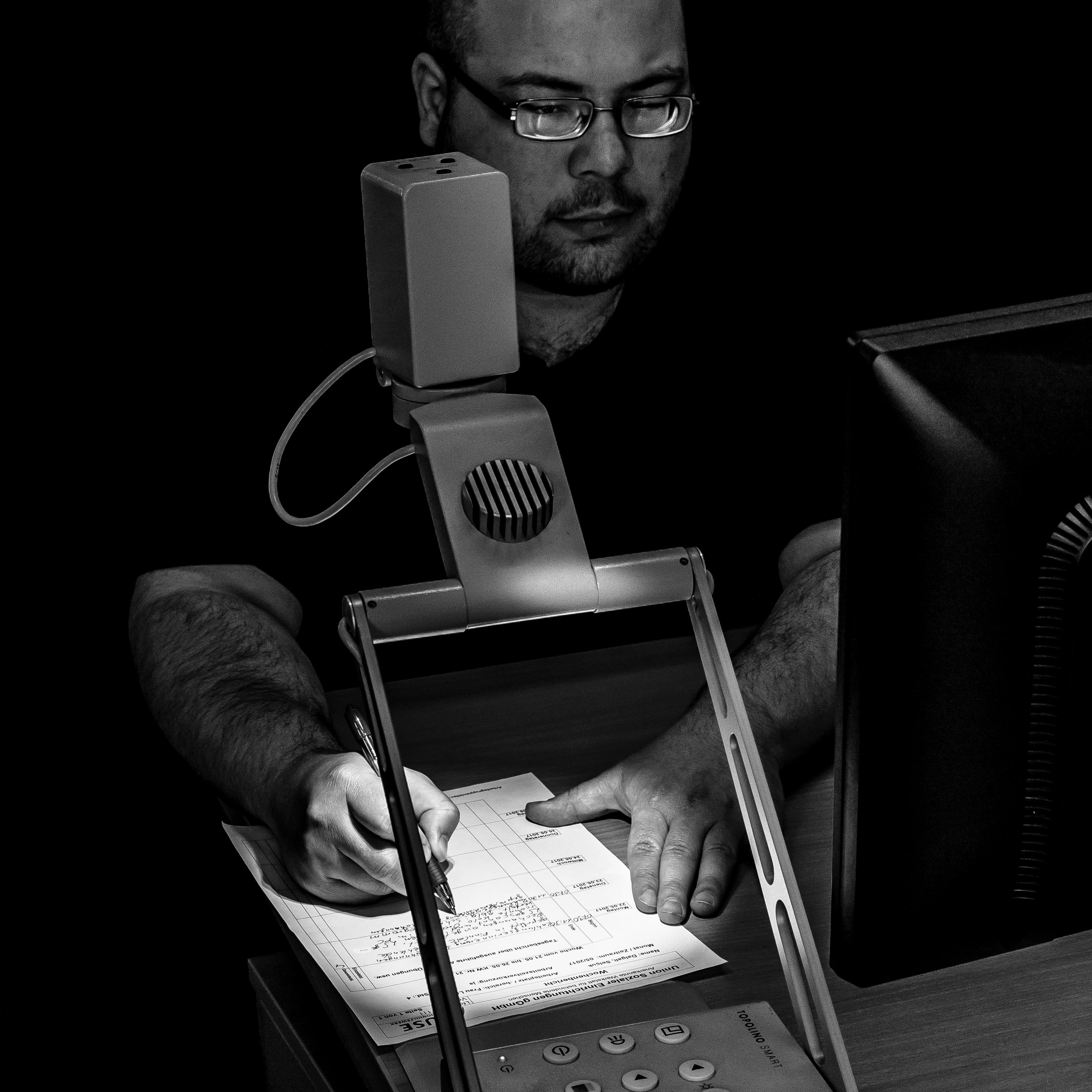 Ein Mann schreibt auf einen Zettel unter einem Lesegerät mit Lampe.