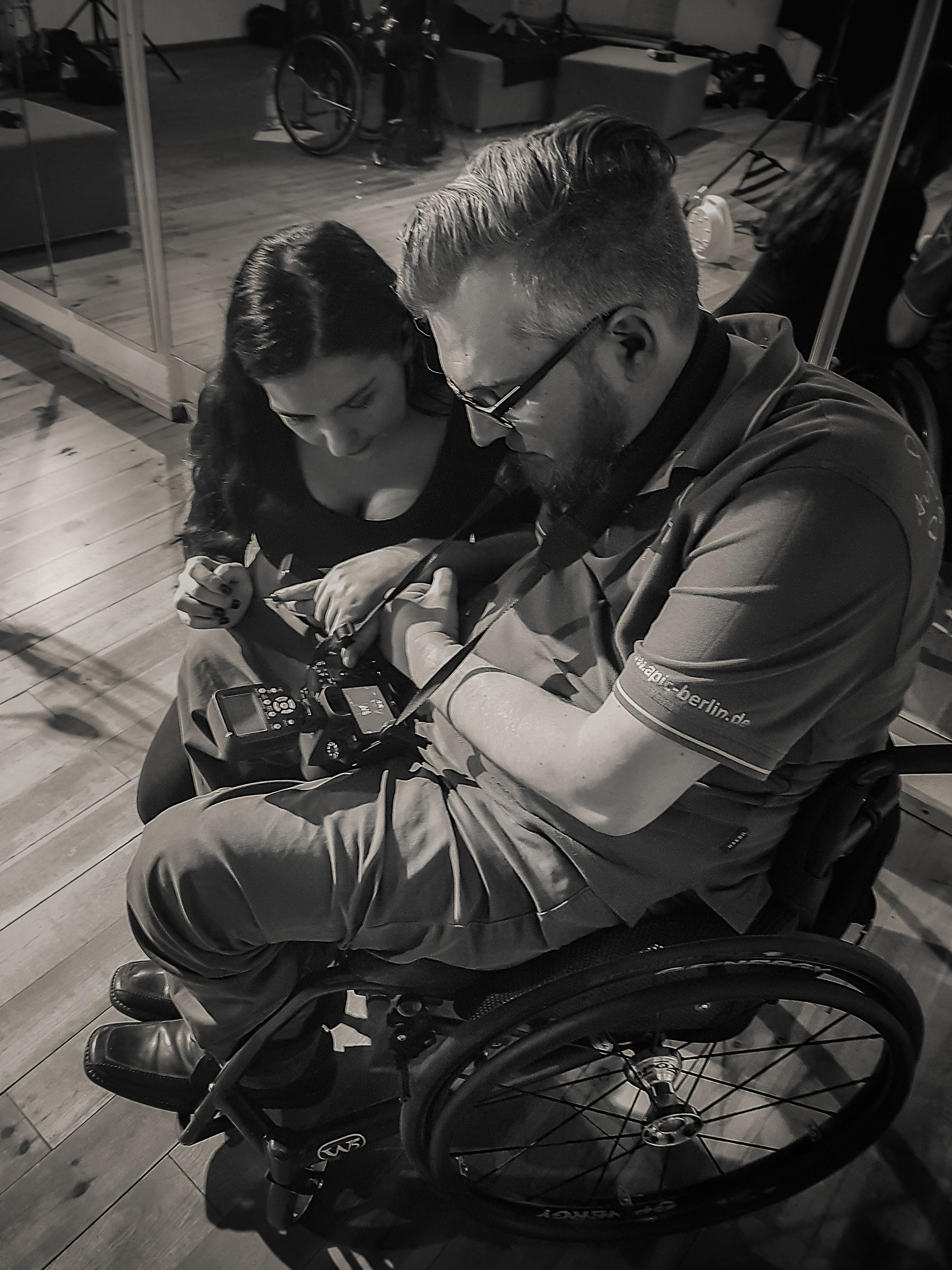 Eine Frau schaut gemeinsam mit einem Fotografen, der im Rollstuhl sitzt , auf das Display einer Fotokamera.