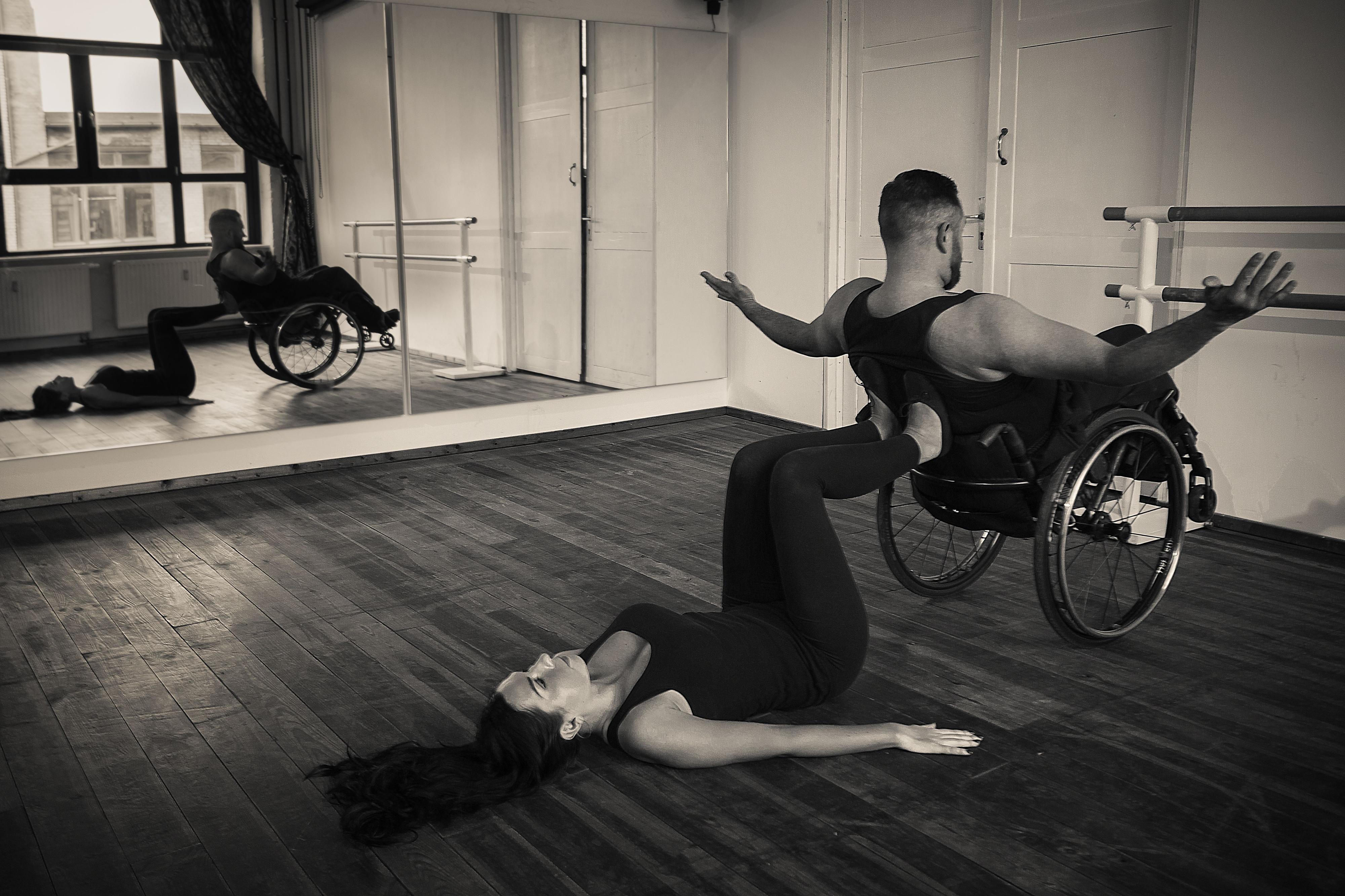 Eine liegt auf dem Boden eines Tanzsaals und stützt mit ihren Füßen einen schräg stehende Rollstuhl, in dem ein Mann sitzt.