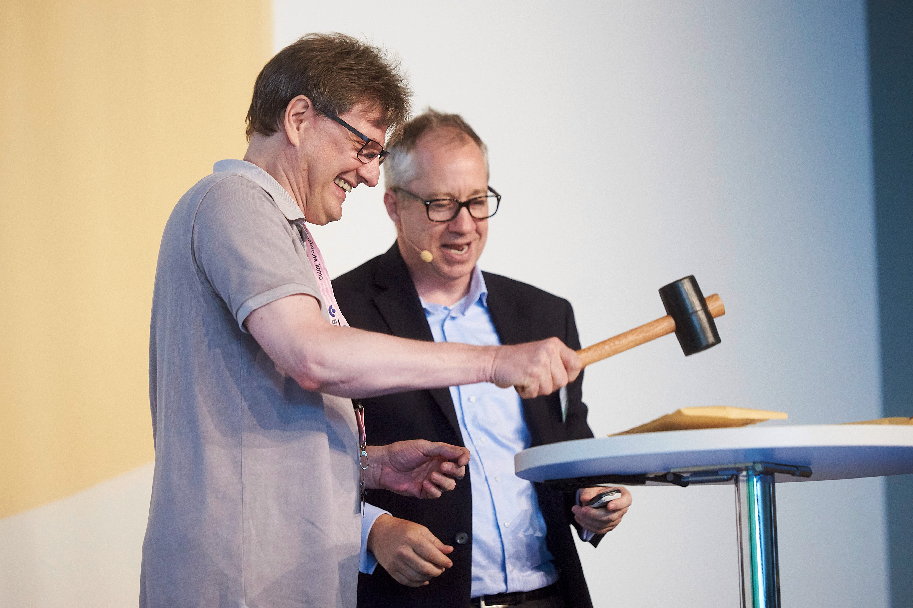 Zwei Männer an einem Stehtisch, auf dem ein Briefumschlag liegt. Einer schlägt mit dem Hammer auf einen Umschlag. Der andere sieht zu. Beide lachen.
