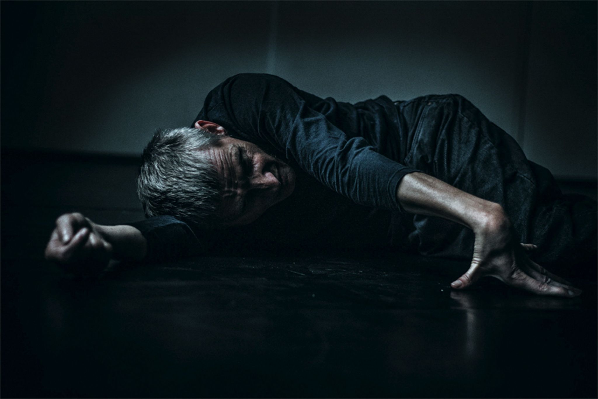 Ein spastisch gelähmter Mann liegt auf der Seite auf dem Boden eines dunklen Zimmers.